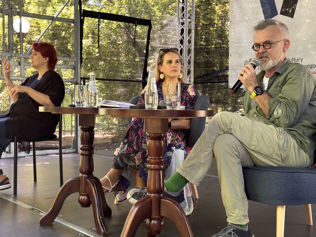 Trzy osoby siedzą na fotelach na scenie w parku. Po lewej stronie siedzi kobieta w czerwonych włosach. Gestykuluje. W centralnej części siedzi kobieta w długich blond włosach. Ma na sobie kwiecistą sukienkę. Patrzy na mężczyznę po prawej stronie. Mężczyzna ubrany w zieloną koszulę, zielone spodnie i zielone skarpetki. Ma okulary. Trzyma w ręce mikrofon. Mężczyzna ma siwe włosy i siwą brodę. Oboje siedzą na fotelach. Przed nimi niskie okrągłe stoliki. Na stolikach szklanki i szklane butelki wody. W tle metalowa konstrukcja.