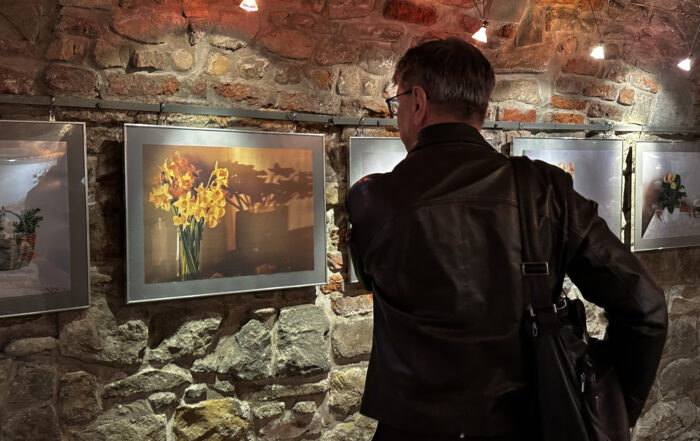 Klimatyczne pomieszczenie z cegieł. Na ścianie obrazy przedstawiające kwiaty. Obrazy oświetlają lampki wystawiennicze. na froncie stoi mężczyzna z przewieszoną na prawym ramieniu torbą i ogląda zdjęcia.