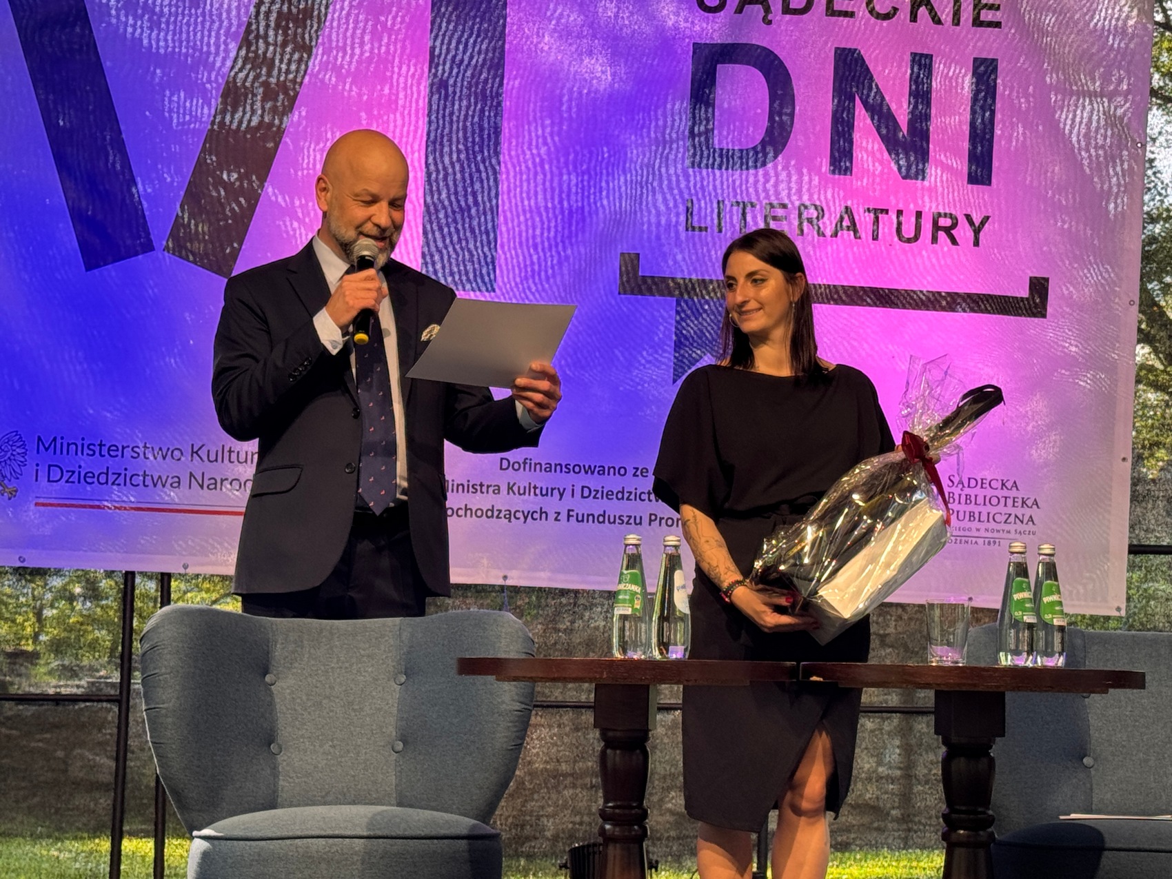 Wręczenie nagród_1 Mężczyzna w ciemnym garniturze i krawacie. Trzyma mikrofon i białą kartkę w ręce. Obok stoi uśmiechnięta kobieta w czarnej sukience. Trzyma prezent w ręce. Przed nimi stoi szary niski fotel i drewniany stół. Na stoliku kilka szklanych butelek. w Tle niebiesko-fioletowy baner.