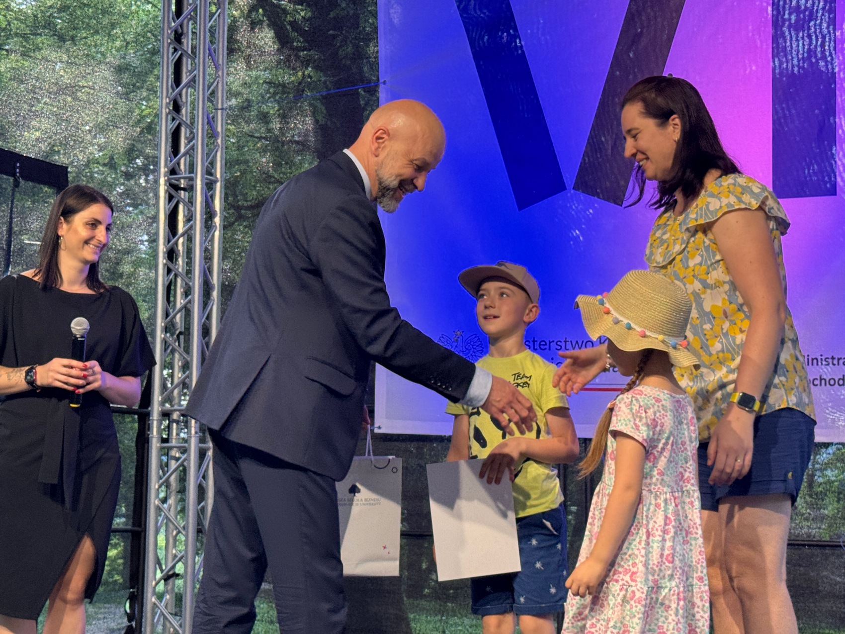 Wręczenie nagród_2 Mężczyzna w garniturze gratuluje kobiecie z dwójką dzieci. Dziewczynka w słomianym kapeluszu w w kwiecistej sukience. Obok chłopiec w żółtej koszulce i czapeczce z daszkiem. Po lewej kobieta w krótkich ciemnych włosach, czarnej sukience. Uśmiecha się i trzyma mikrofon w ręce. w tle niebiesko-fioletowy baner.