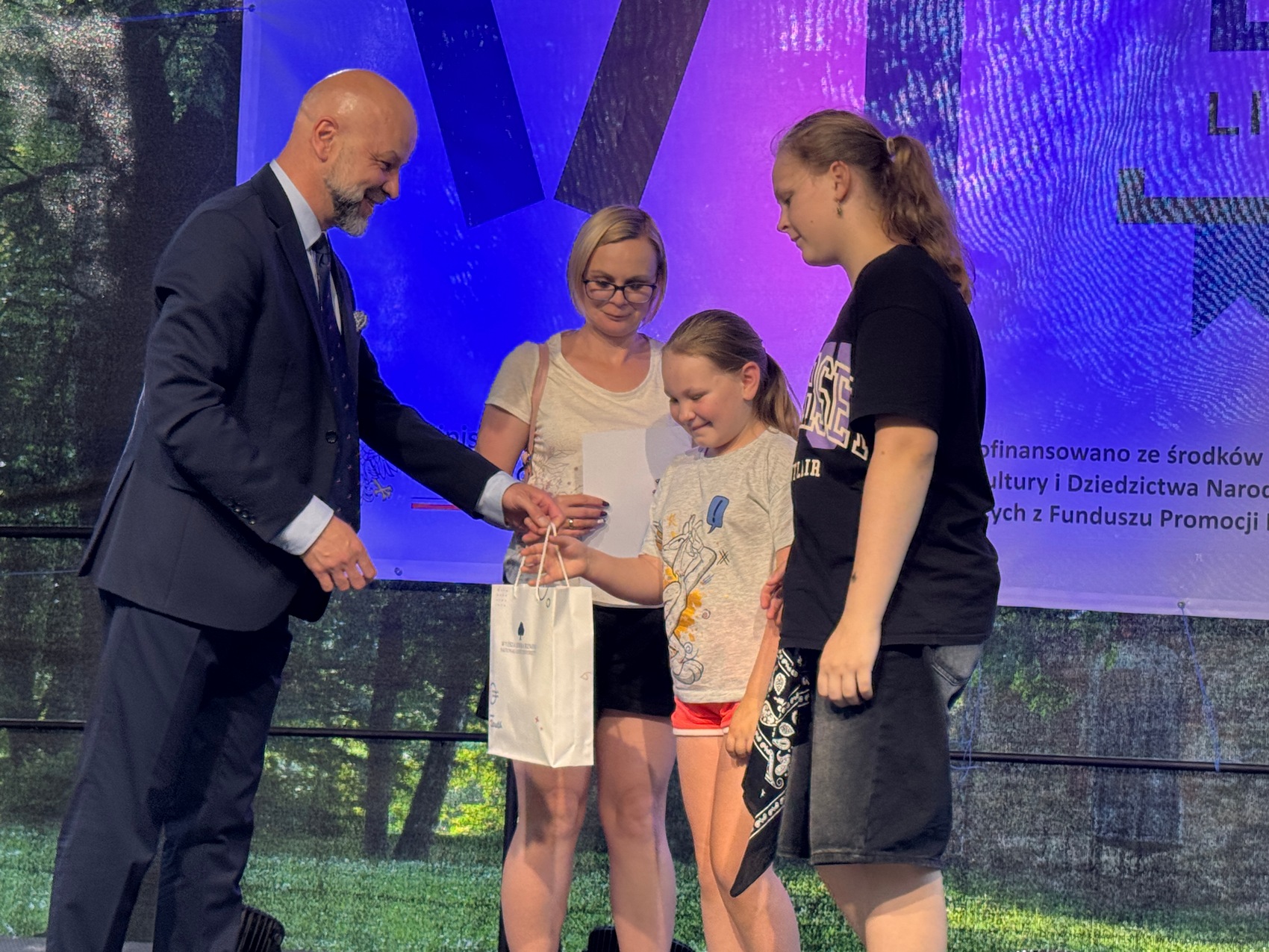Wręczenie nagród_3 Mężczyzna w ciemnym garniturze podaje białą torebkę dziewczynce. Obok niej stoją dwie kobiety. Dziewczyna po prawej stronie dziewczyna w kucyku, czarnej koszulce i krótkich spodenkach. W tle niebiesko-fioletowy baner.