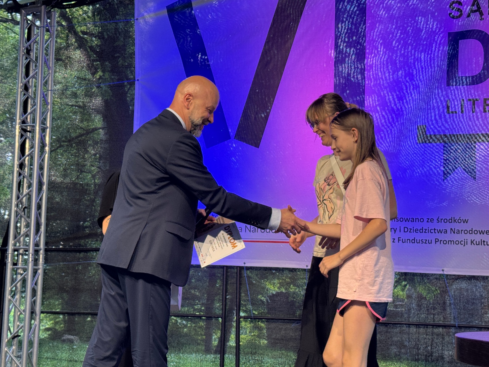 Wręczenie nagród_4 Mężczyzna w ciemnym garniturze gratuluje dwóm kobietom. Uśmiechają się. Dziewczyny ubrane w jasne podkoszulki. W tle baner niebiesko-fioletowy.