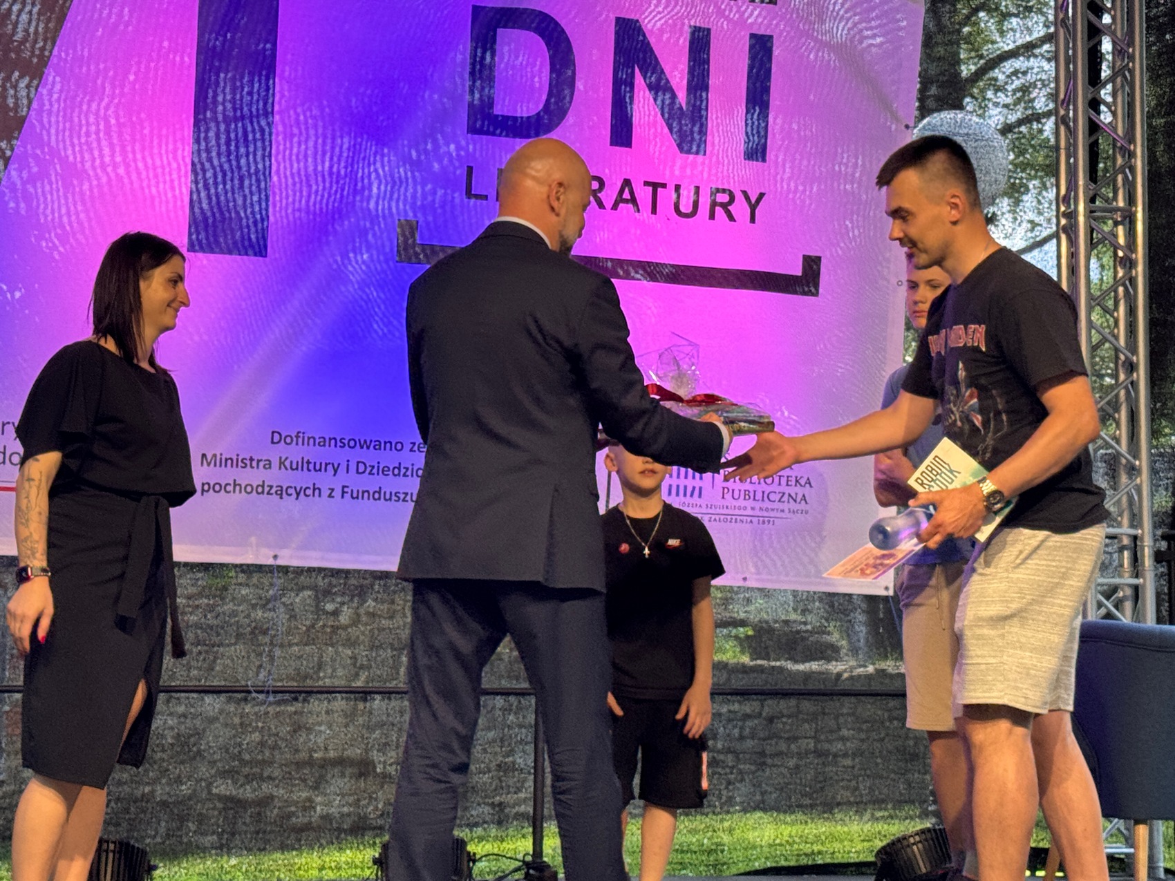 Wręczenie nagród_5 Pięć osób na scenie. W centralnym miejscu mężczyzna w ciemnym garniturze przekazuje płaski zapakowany prezent. Mężczyzna po prawej stronie odbiera nagrodę. Ubrany jest w ciemną koszulkę i jasne spodnie. Za nimi stoi dwójka chłopców. W tle niebiesko-fioletowy baner. Po lewej stronie stoi dziewczyna w krótkich czarnych włosach i w czarnej sukience.