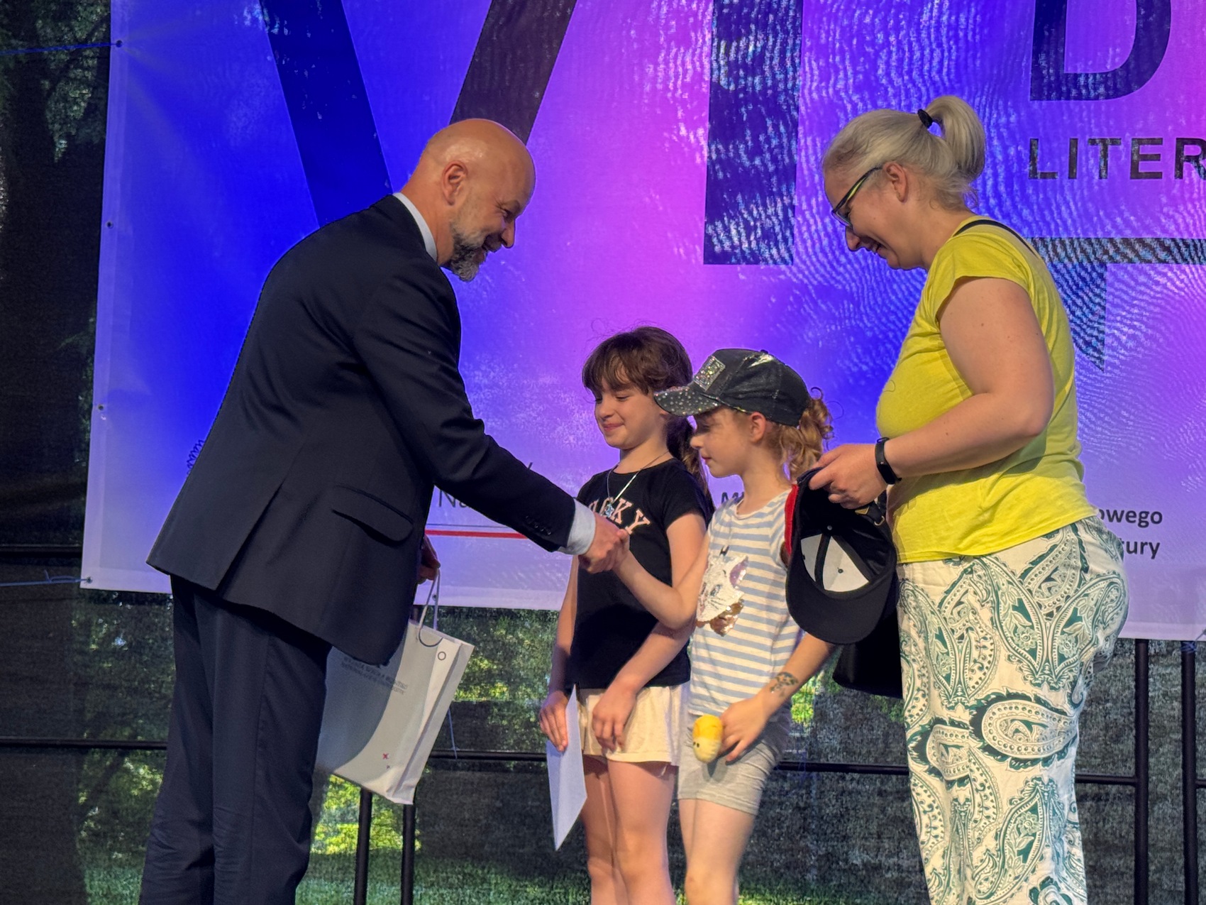 Wręczenie nagród_6 Cztery osoby. Po lewej łysy mężczyzna w czarnym garniturze gratuluje dwóm małym dziewczynkom na scenie. Obok nich stoi kobieta w jasnym kucyku. Ma na sobie żółtą bluzkę i spodnie w jasnoniebieskie wzorki. Za nimi duży niebiesko-fioletowy baner.