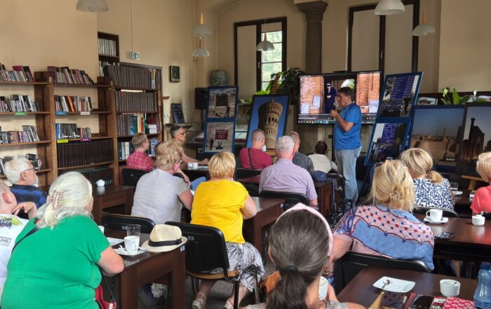 Sala wypełniona publiką. Ludzie siedzą przy ciemnobrązowych prostokątnych stołach. Zwróceni są tyłem do obiektywu. Patrzą na duży ekran na froncie i kolorowe obrazy, przy których stoi mężczyzna w niebieskiej koszulce sportowej. Mężczyzna mówi do mikrofony. Po lewej stronie na ścianie duży regał z książkami.
