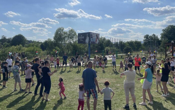 Plener. Na łące stoi w okręgu kilkadziesiąt osób; dzieci, kobiet i mężczyzn. W tle duży bilbord z reklamą w ciemnych kolorach.