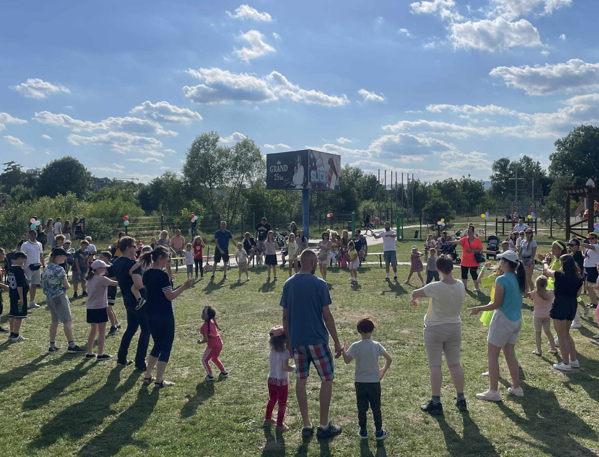 Plener. Na łące stoi w okręgu kilkadziesiąt osób; dzieci, kobiet i mężczyzn. W tle duży bilbord z reklamą w ciemnych kolorach.