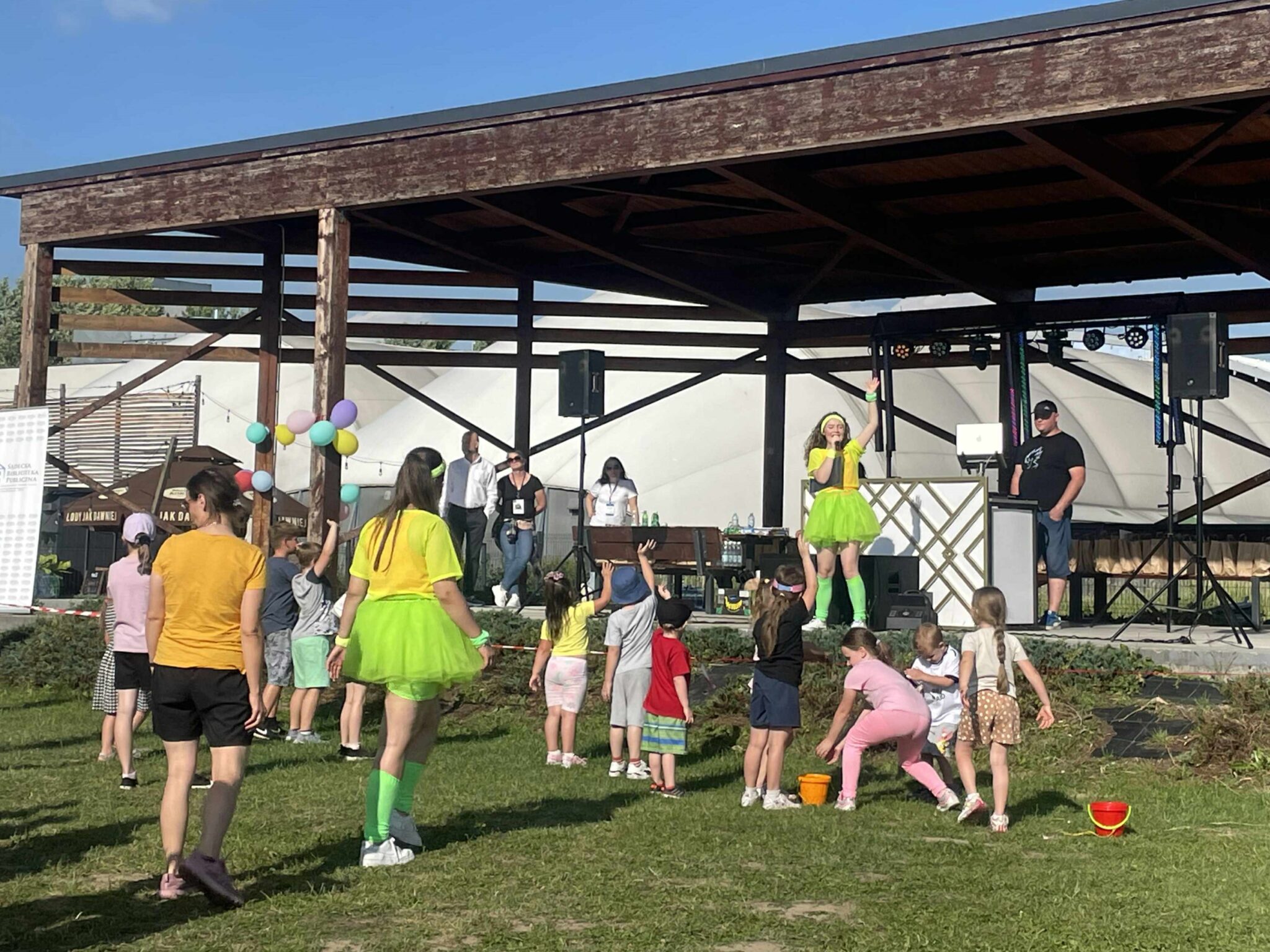 Piknik_disco_budżet_3 Łąka z drewnianą wiatą. Pod wiatą 5 osób. Jedna z nich - kobieta - ubrana jest w odblaskową żółtą koszulkę oraz odblaskową jasnozieloną krótką spódniczkę i podkolanówki. Ma podniesioną lewą rękę i zwrócona jest do grupy kilkunastu osób, które stoją na łące i patrzą na nią.