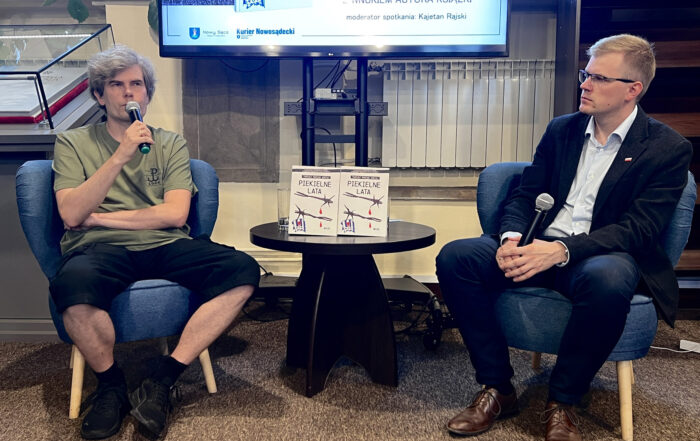 Dwóch mężczyzn siedzi w sali na niskich niebieskich fotelach. Mężczyzna z lewej ma gęste siwe włosy, zieloną koszulkę i krótkie czarne spodenki. Trzyma w ręce mikrofon i trzyma go przy twarzy. Po prawej siedzi drugi z mężczyzn. Ma na sobie ciemny garnitur i jasną koszulę. Patrzy w skupieniu na mężczyznę obok. Trzyma w ręce mikrofon. Pomiędzy nimi niski okrągły stolik. Na stoliku stoją dwie książki. Nad nimi fragment dużego ekranu.