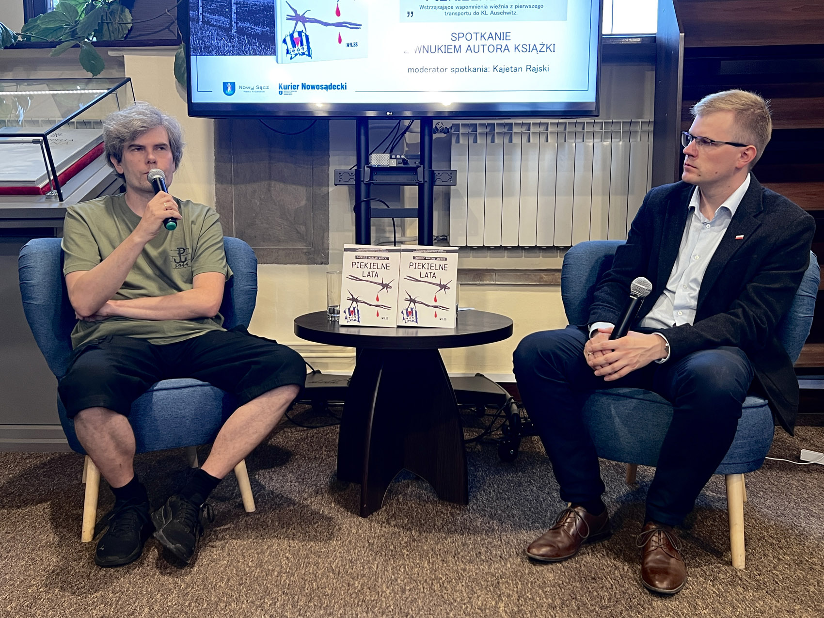 Dwóch mężczyzn siedzi w sali na niskich niebieskich fotelach. Mężczyzna z lewej ma gęste siwe włosy, zieloną koszulkę i krótkie czarne spodenki. Trzyma w ręce mikrofon i trzyma go przy twarzy. Po prawej siedzi drugi z mężczyzn. Ma na sobie ciemny garnitur i jasną koszulę. Patrzy w skupieniu na mężczyznę obok. Trzyma w ręce mikrofon. Pomiędzy nimi niski okrągły stolik. Na stoliku stoją dwie książki. Nad nimi fragment dużego ekranu.