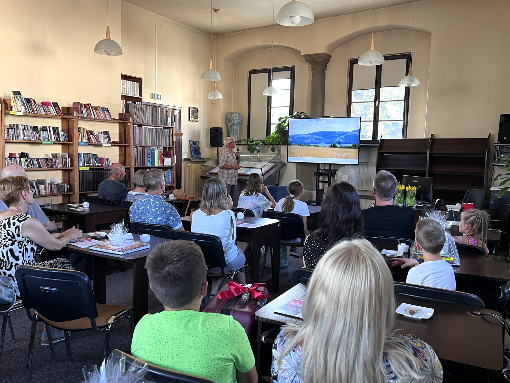 Sądecczyzna na rowerze (2) Sala wypełniona publiką. Ludzie siedzą przy ciemnobrązowych prostokątnych stołach. Zwróceni są tyłem do obiektywu. Patrzą na duży ekran na froncie, przy którym stoi mężczyzna w jasnej koszuli z krótkim rękawem. Mężczyzna mówi do mikrofonu. Po lewej stronie na ścianie duży regał z książkami.