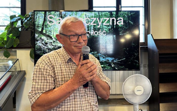 Portret mężczyzny, od pasa w górę. Mężczyzna znajduje się w pomieszczeniu. Ubrany jest w jasną koszulę z krótkim rękawem w kratkę. Na nosie ma okulary. W obydwóch rękach trzyma mikrofon blisko ust. Za mężczyzną duży ekran.