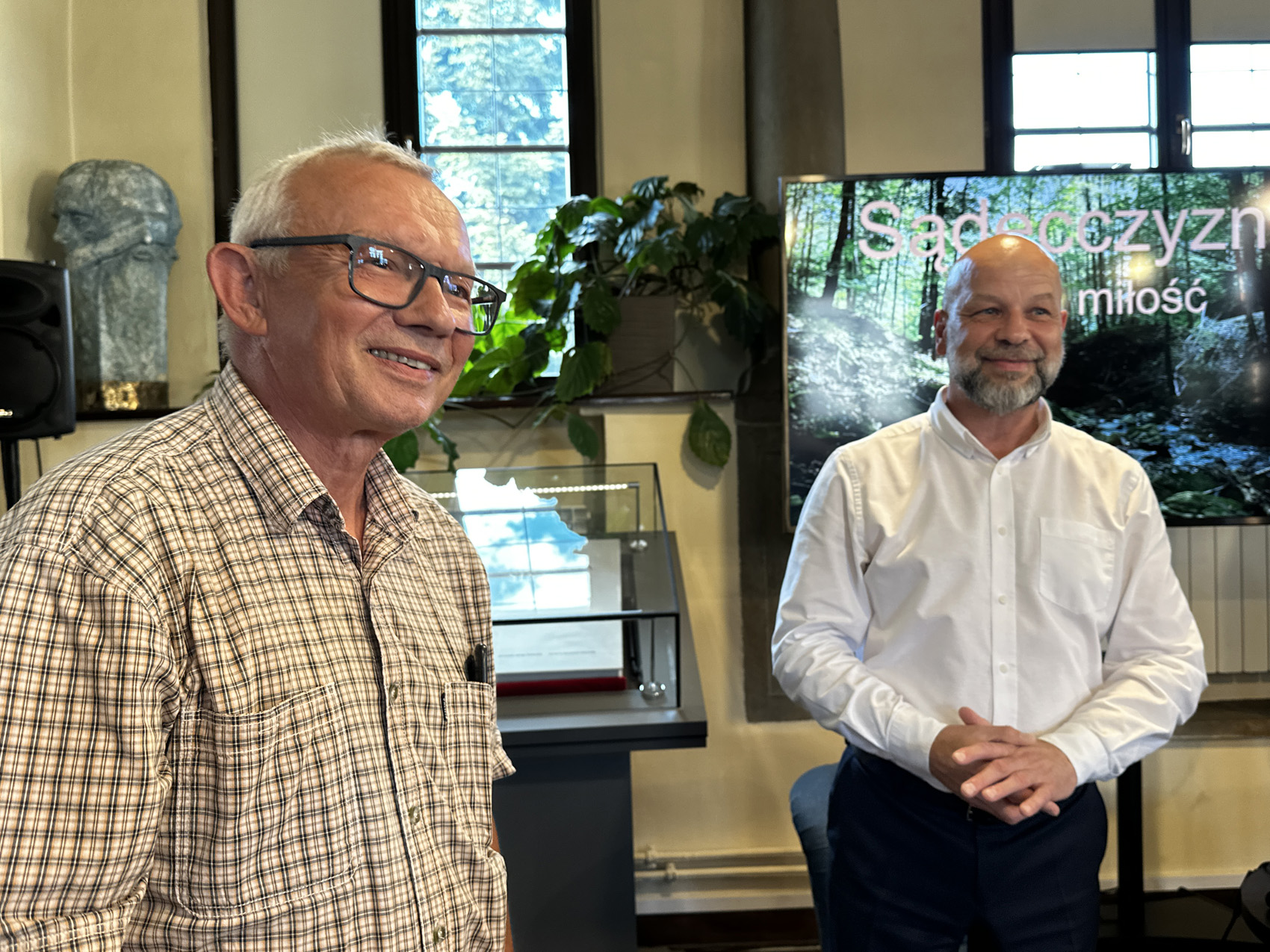 Sądecczyzna na rowerze (4) W pomieszczeniu stoi dwóch mężczyzn. Patrzą przed siebie, uśmiechają się. Mężczyzna po lewej stronie na nosie ma okulary i ubrany jest w jasną koszulę z krótkim rękawem w kratkę. Mężczyzna po prawej stronie ubrany jest w białą elegancką koszulę i granatowe spodnie.