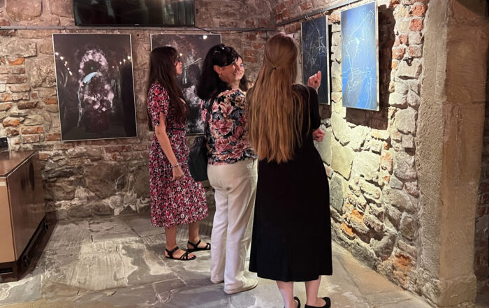 Cztery kobiety. Sala z kamiennymi ścianami. Kobiety patrzą na grafiki wiszące na ścianach. Ubrane są elegancko i stoją tyłem. Uśmiechają się.