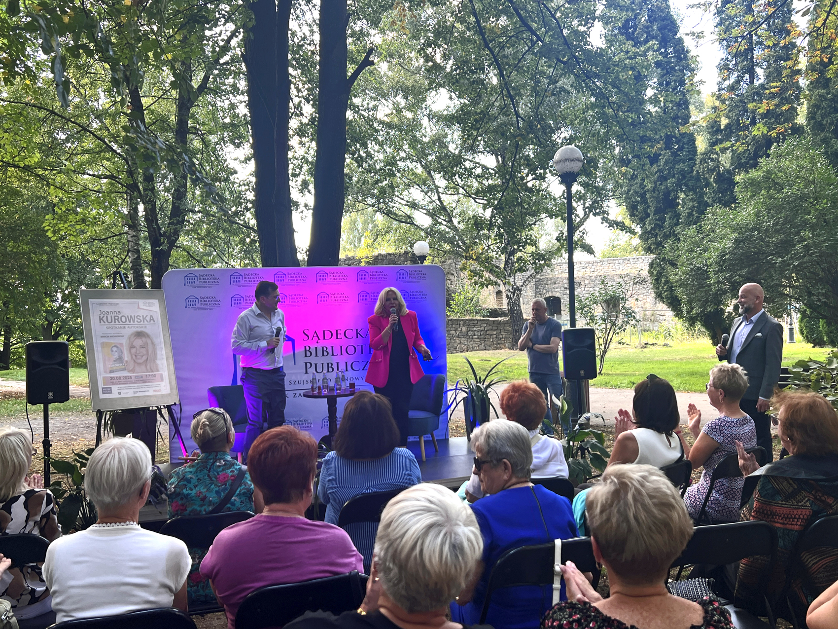 Joanna Kurowska – spotkanie autorskie (1) Scena w parku miejskim, w tle ruiny zamku. Przed sceną publika siedząca na krzesłach patrzy na scenę. Na scenie mężczyzna w białej koszuli i granatowych spodniach oraz kobieta w różowej marynarce. W tle podświetlona na niebiesko-różowo biała ścianka reklamowa z niebieskimi napisami.