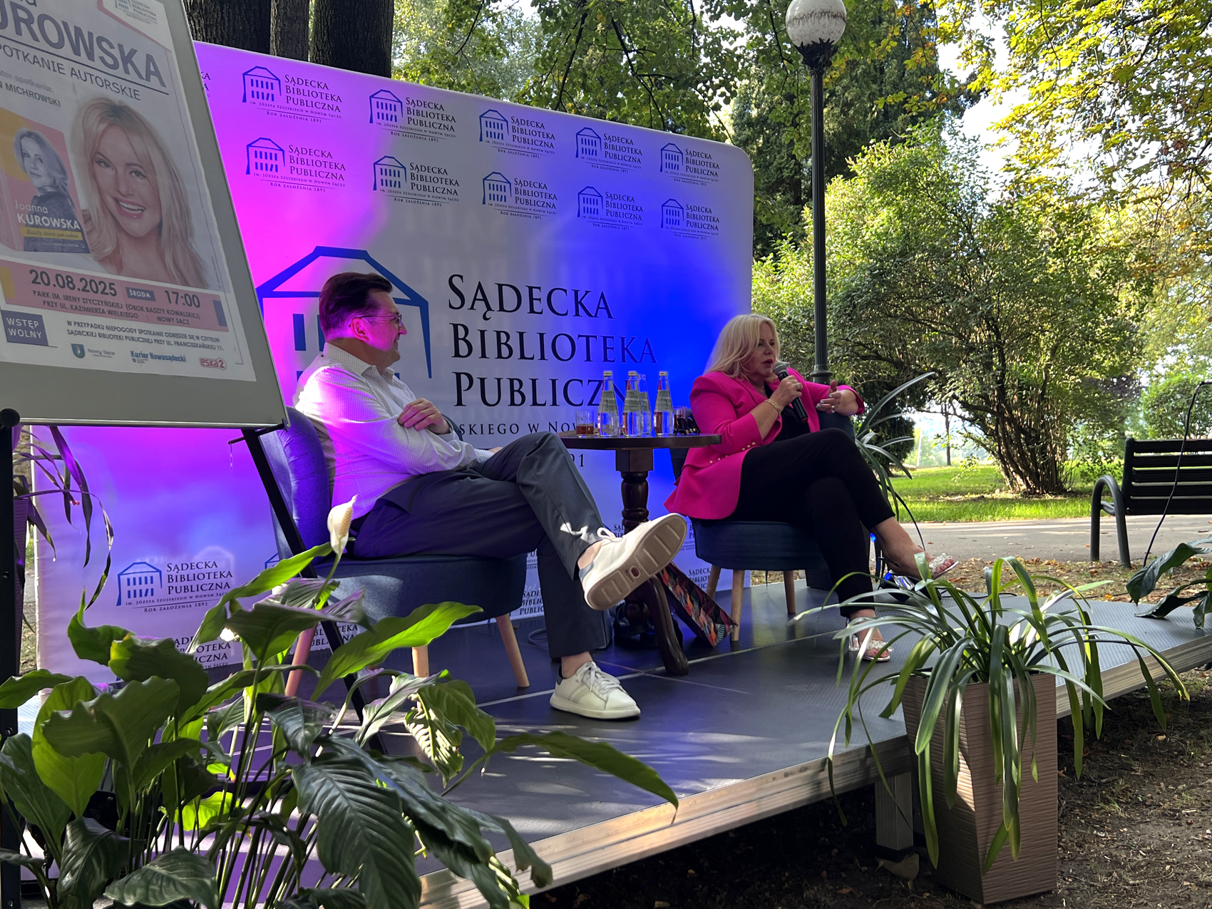 Joanna Kurowska – spotkanie autorskie (2) Scena w parku. Wokół zielone drzewa. Na scenie siedzi mężczyzna w białej koszuli i granatowych spodniach. oraz kobieta z długimi blond włosami. Kobieta ma na sobie czarne spodnie i różową marynarkę. W tle podświetlona na różowo-niebiesko ścianka reklamowa.