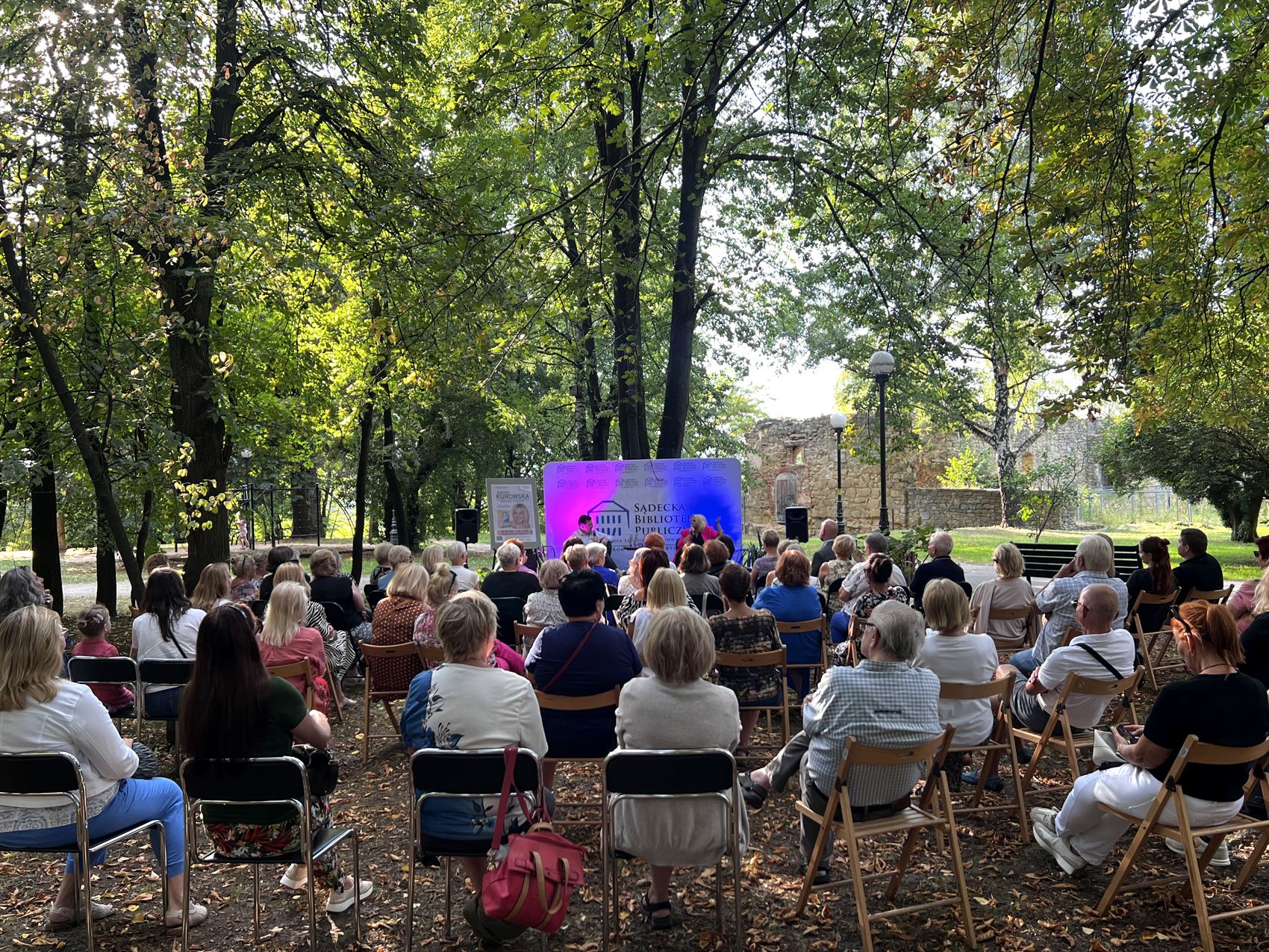 Joanna Kurowska – spotkanie autorskie (3) Park, w tle ruiny zamku. Widownia licząca kilkadziesiąt osób siedzi zwrócone w stronę podświetlonej na biało-różowo ścianki. Przed ścianką siedzą mężczyzna i kobieta.