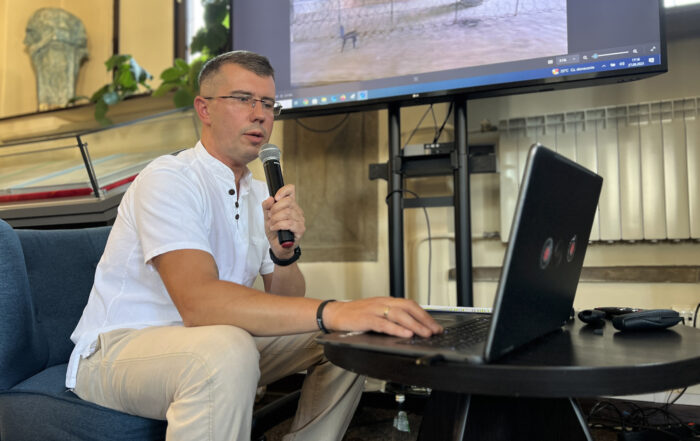 Mężczyzna w średnim wieku w okularach siedzi na niebieskim fotelu przy okrągłym ciemnobrązowym stoliku. W lewej ręce ma mikrofon, prawą pisze na klawiaturze laptopa. Za nim duży ekran z kolorową grafiką.