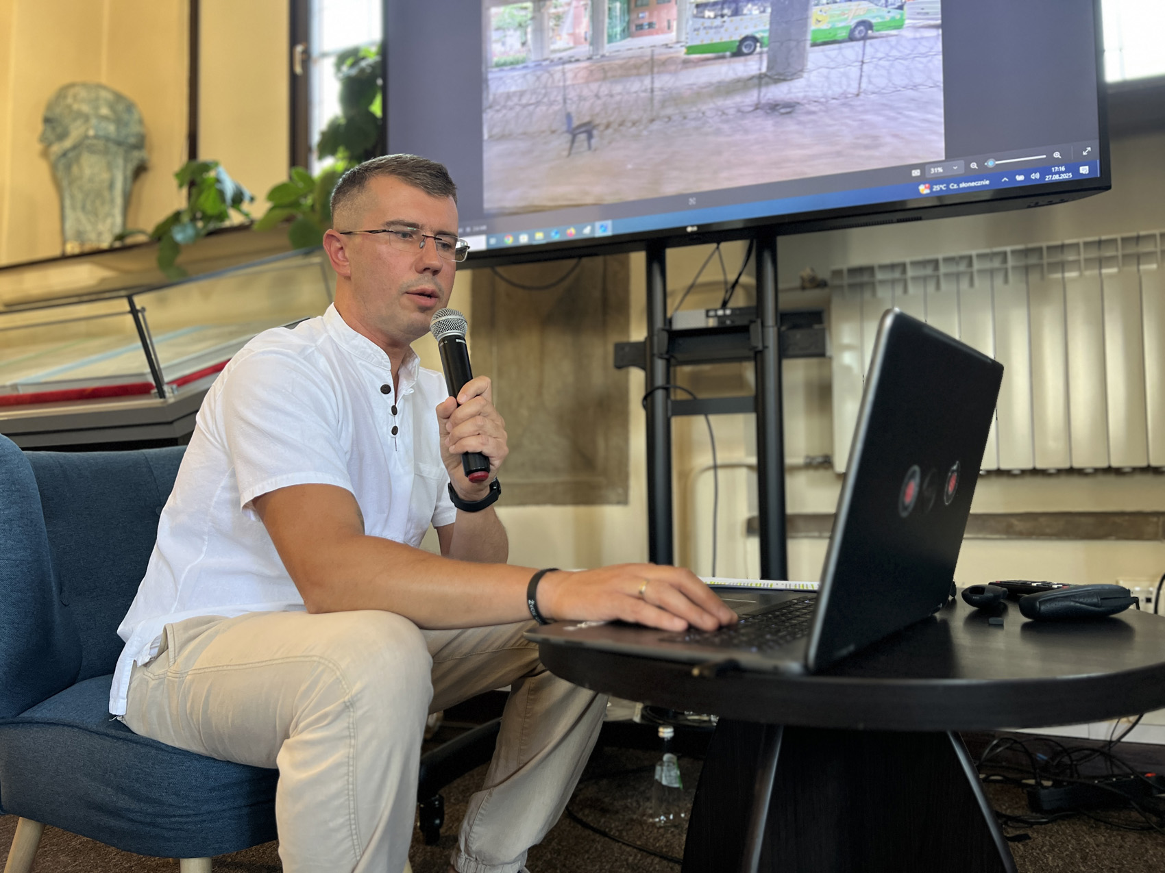 Mężczyzna w średnim wieku w okularach siedzi na niebieskim fotelu przy okrągłym ciemnobrązowym stoliku. W lewej ręce ma mikrofon, prawą pisze na klawiaturze laptopa. Za nim duży ekran z kolorową grafiką.