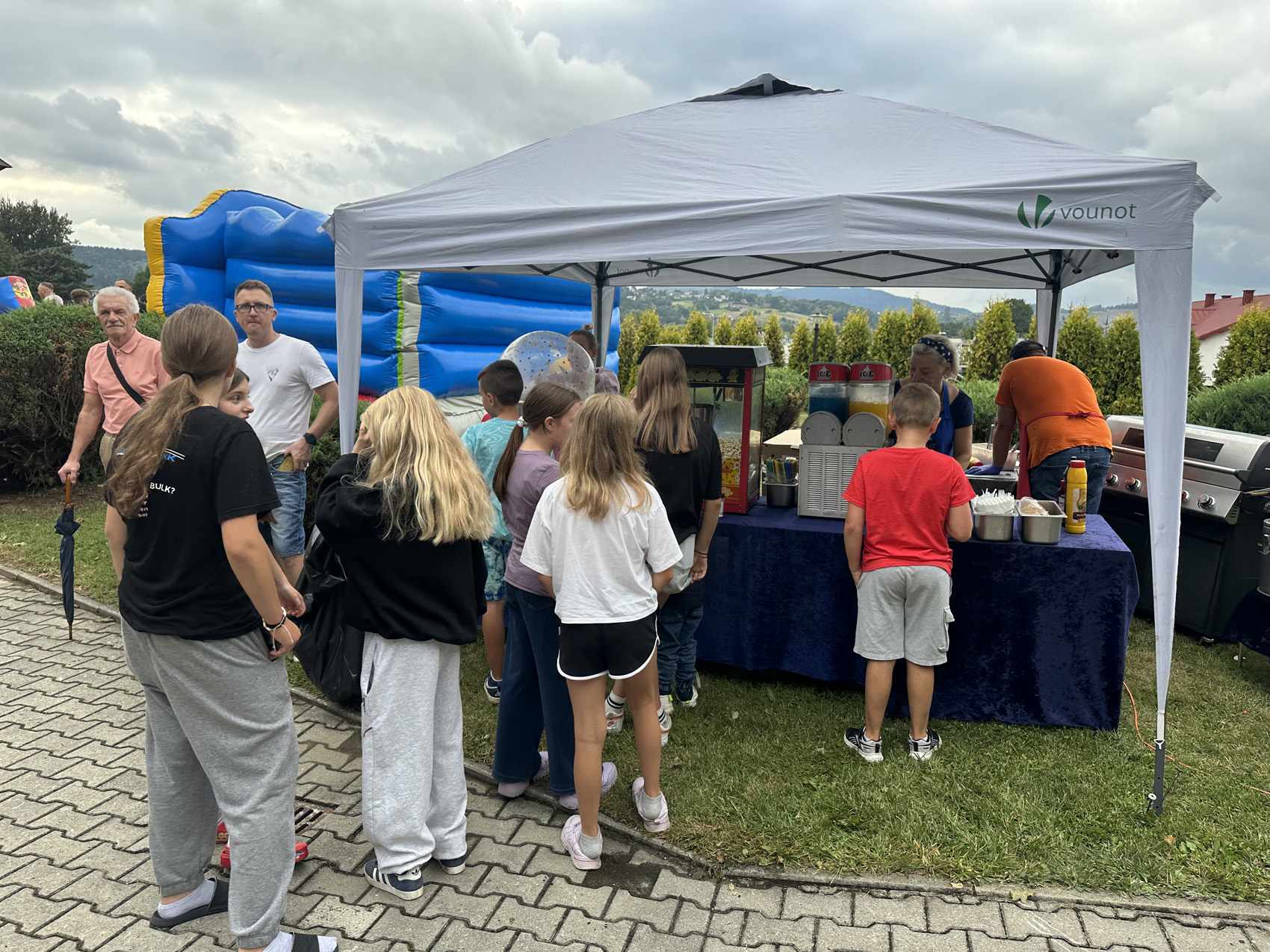 Festyn Zabełcze – Budżet Obywatelski (1) Plener. Na środku stoi szary namiot. Pod namiotem stoi . stół nakryty granatowym długim obrusem. Na stole stoją: maszyna z popcornem, napoje, sztućce Za stołem stoi kobieta i mężczyzna. Przed stołem stoi kolejka ludzi: młodzieży i dorosłych. W tle widać niebieski dmuchany namiot dla dzieci.