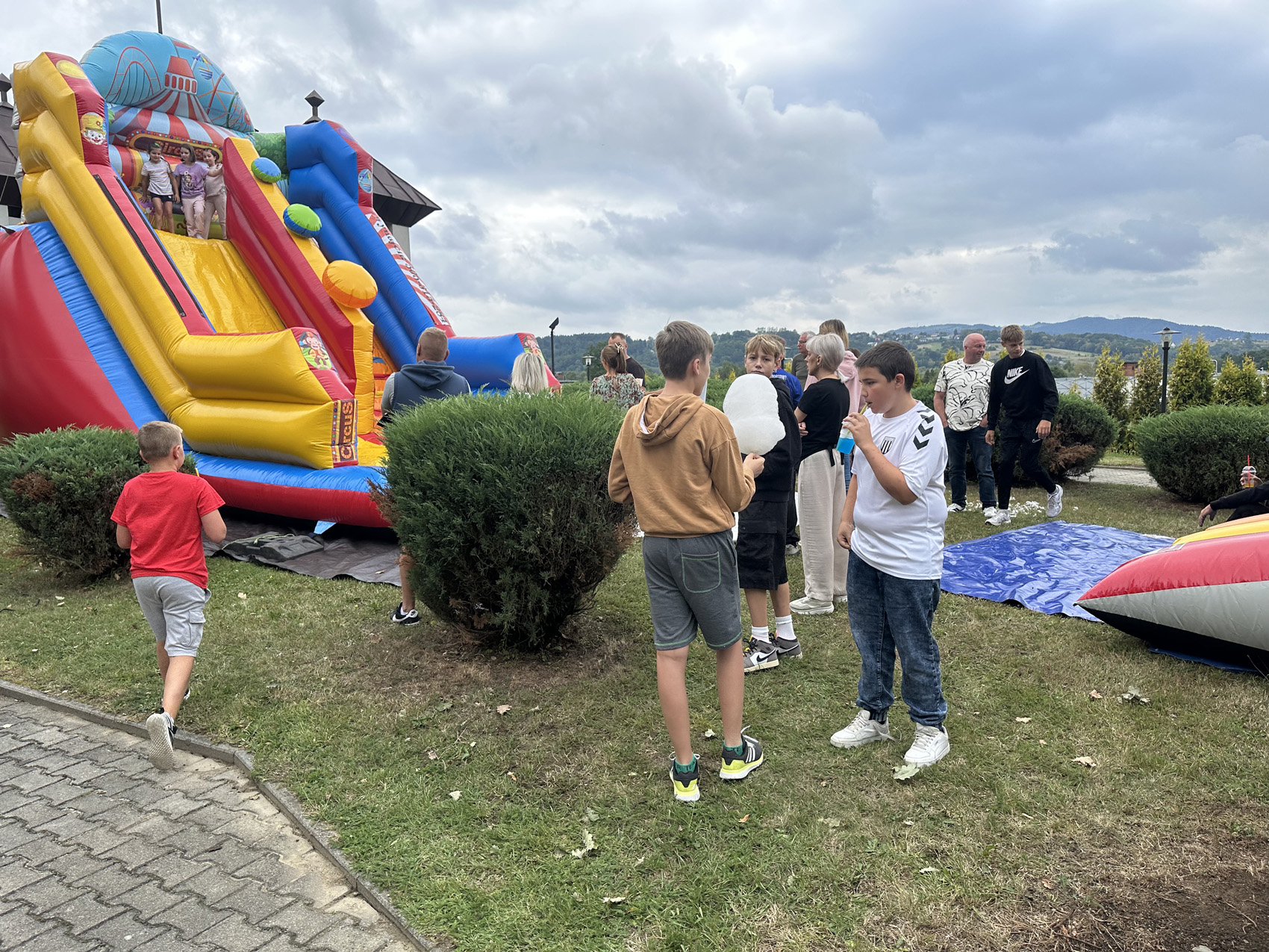 Festyn Zabełcze – Budżet Obywatelski (2) Plener. Na środku na trawie stoi grupa nastolatków. Rozmawiają ze sobą. Po lewej stronie stoi duża kolorowa dmuchana zjeżdżalnia. Na szczycie zjeżdżalni stoją trzy młode dziewczynki.