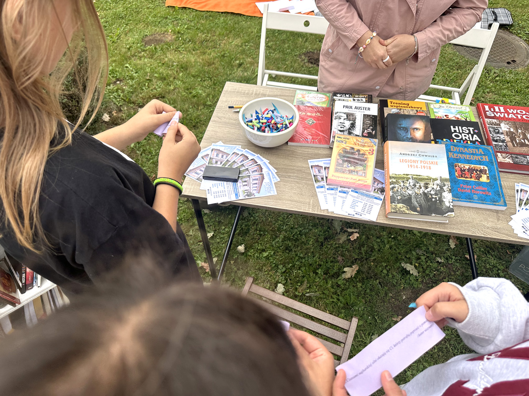 Festyn Zabełcze – Budżet Obywatelski (3) Plener. Na środku stoi długi stolik. Na stoliku rozłożone są książki z kolorowymi okładkami. Obok książki po lewej stronie stoi biała miseczka z kolorowymi karteczkami. Za stolikiem stoi kobieta w różowym płaszczu pokazana od łokci w dół. W dolnym kadrze pokazane od góry trzy młode dziewczyny. W rękach trzymają kolorowe karteczki z napisem.