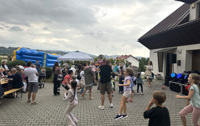 Otwarta przestrzeń na świeżym powietrzu. Na placu chodnikowym stoi grupa osób: dorośli i dzieci. Pokazani są od tyłu i z boku. Dzieci na środku tańczą. Otwarta przestrzeń na świeżym powietrzu. Na placu chodnikowym stoi grupa osób: dorośli i dzieci. Pokazani są od tyłu. Dzieci tańczą. Otwarta przestrzeń na świeżym powietrzu. Na placu chodnikowym stoi grupa osób: dorośli i dzieci. Pokazani są od tyłu. Dzieci tańczą. Po prawej stronie widać kawałek budynku z białą elewacją i brązowym dachem. W powietrzu unoszą się duże kolorowe bańki mydlane. Przed bramą garażową budynku stoi duża czarna skrzynia i żółte światła. W powietrzu unoszą się duże kolorowe bańki mydlane. Za grupą osób stoi budynek z białą elewacją i brązowym dachem. W powietrzu unoszą się duże kolorowe bańki mydlane.