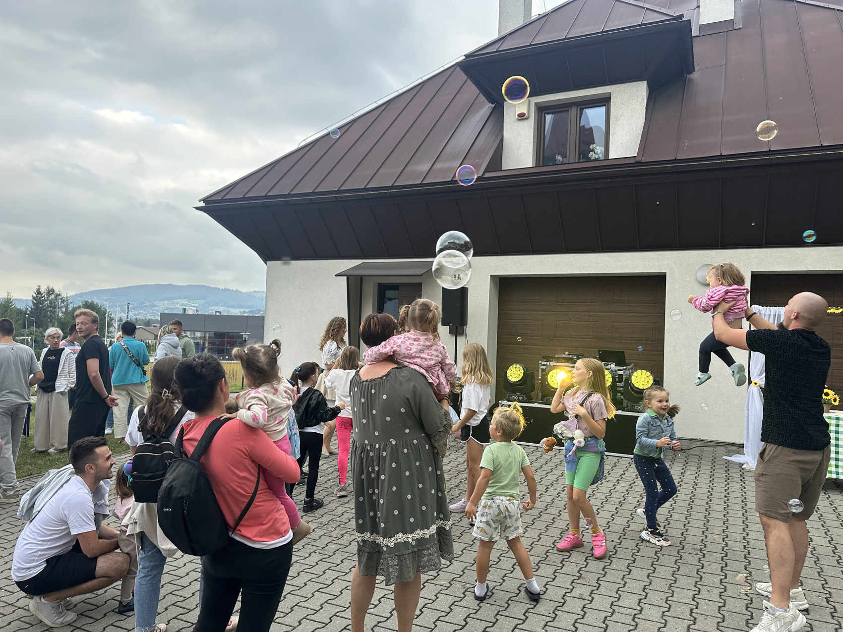 Festyn Zabełcze – Budżet Obywatelski (5) Otwarta przestrzeń na świeżym powietrzu. Na placu chodnikowym stoi grupa osób: dorośli i dzieci. Pokazani są od tyłu. Dzieci tańczą. Za grupą osób stoi budynek z białą elewacją i brązowym dachem. Przed bramą garażową budynku stoi duża czarna skrzynia i żółte światła. W powietrzu unoszą się duże kolorowe bańki mydlane.