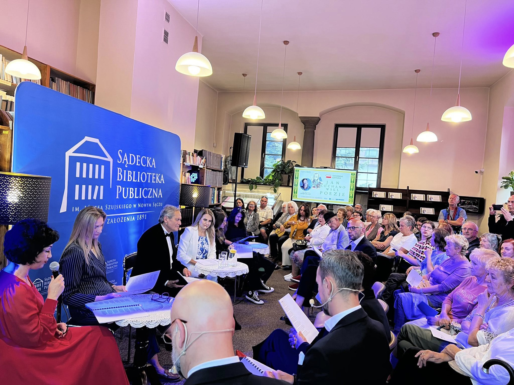 Sala wypełniona siedzącymi na krzesłach osobami. Widownia patrzy na siedzących przy trzech stolikach sześć ubranych na galowo osób. W tle duży ekran z kolorową grafiką.