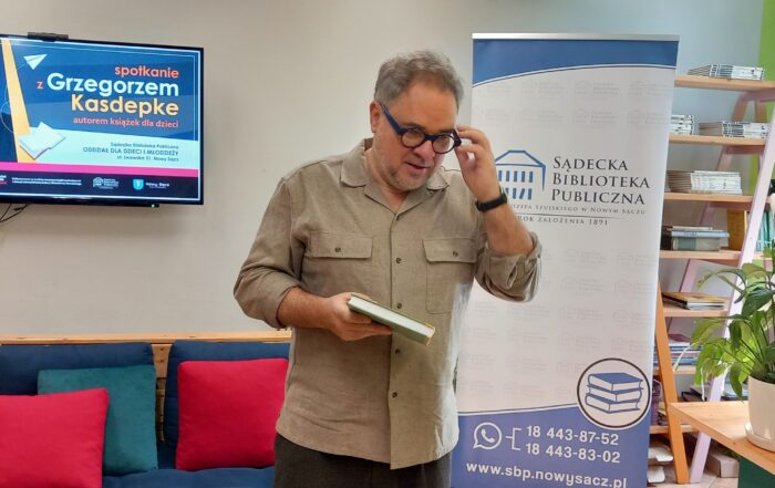 Wnętrze czytelni. Pośrodku stoi mężczyzna, w ręce trzyma książkę, drugą zakłąda okulary. W tle ekran z napisem "spotkanie z Grzegorzem Kasdepke" oraz baner z logo Sądeckiej Biblioteki Publicznej.