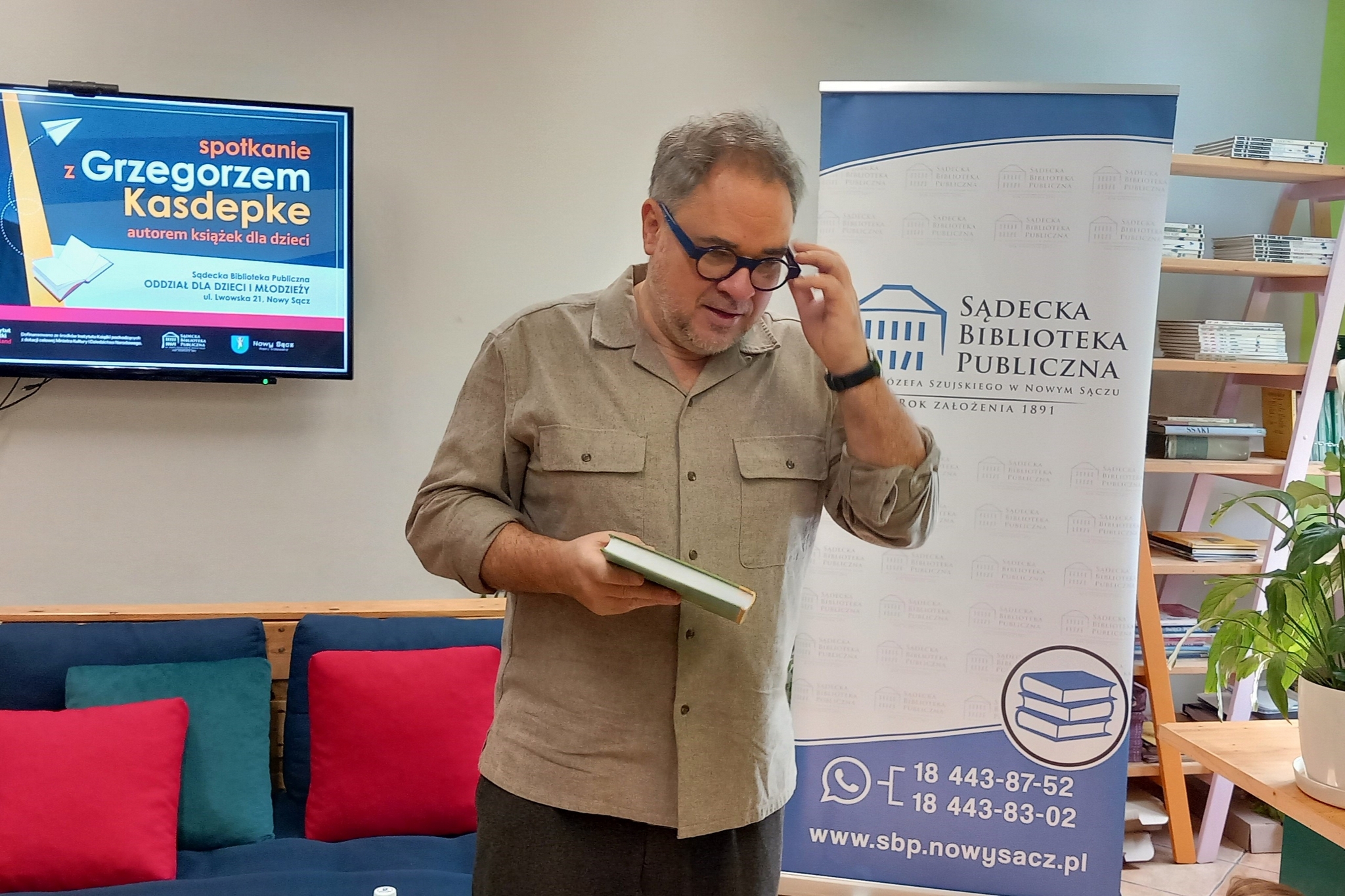 Wnętrze czytelni. Pośrodku stoi mężczyzna, w ręce trzyma książkę, drugą zakłąda okulary. W tle ekran z napisem "spotkanie z Grzegorzem Kasdepke" oraz baner z logo Sądeckiej Biblioteki Publicznej.