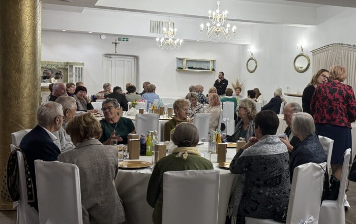 Na zdjęciu widać salę bankietową, w której odbywa się uroczystość. W pomieszczeniu znajduje się wiele osób siedzących przy okrągłych stołach, nakrytych białymi obrusami. Krzesła również są białe i ozdobione kokardami. Na stołach stoją talerze, szklanki i butelki z napojami. W tle widać dekoracje, takie jak żyrandole, lustra i obrazy. Na ścianie wisi krzyż.
