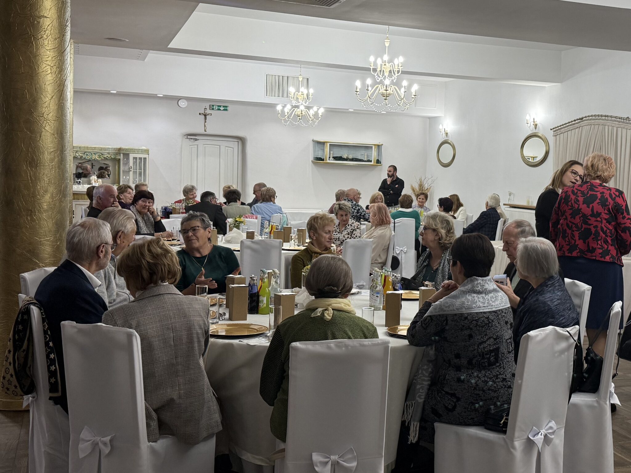 Na zdjęciu widać salę bankietową, w której odbywa się uroczystość. W pomieszczeniu znajduje się wiele osób siedzących przy okrągłych stołach, nakrytych białymi obrusami. Krzesła również są białe i ozdobione kokardami. Na stołach stoją talerze, szklanki i butelki z napojami. W tle widać dekoracje, takie jak żyrandole, lustra i obrazy. Na ścianie wisi krzyż.