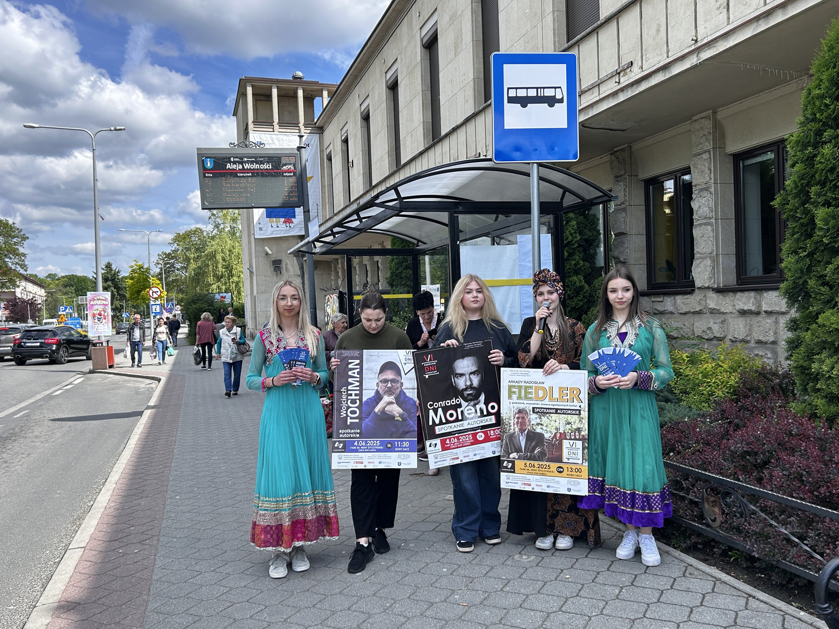Happening młodzieżowy – SDL (1) Pięć młodych dziewczyn stoi przed dużym budynkiem i przystankiem autobusowym. Dwie z nich są ubrane w niebieskie suknie. Jedna z nich ma złoto-różowe wstawki, a druga złoto-fioletowe. Obie trzymają zakładki. Pomiędzy nimi stoją trzy inne dziewczyny