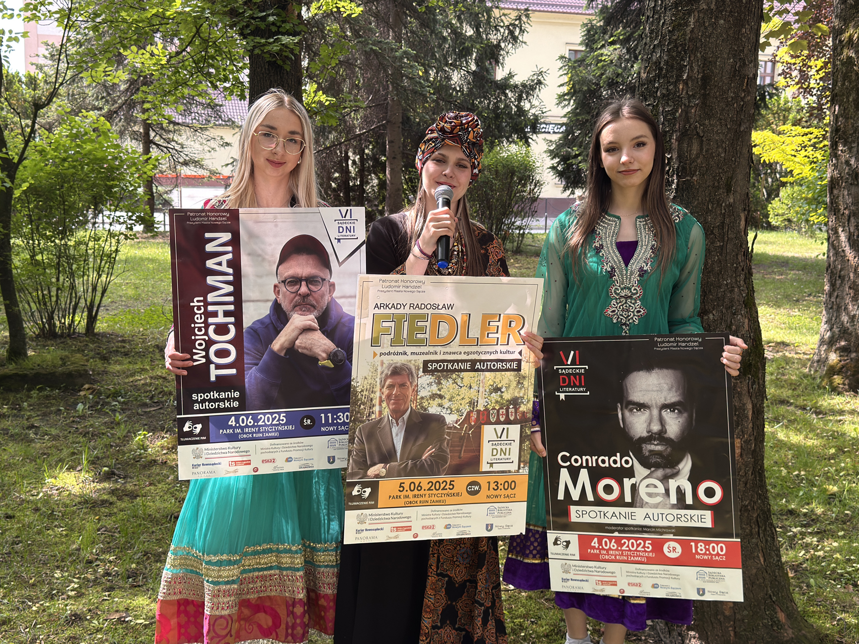 Happening młodzieżowy – SDL (2) Trzy młode dziewczyny stoją w parku. Pierwsza z nich po lewej ma blond włosy i okulary. Ubrana jest w długą zieloną sukienkę ze złoto-różowymi wstawkami trzyma w rękach plakat zapowiadający spotkanie z Wojciechem Tochmanem. Obok niej stoi dziewczyna w czarnej długiej sukience w pomarańczowo-czerwone wzory. Na głowie ma pasujący turban. W ręce trzyma mikrofon. W ręce trzyma plakat zapowiadający spotkanie z Arkadym Radosławem Fiedlerem. Po prawej stoi trzecia z dziewczyn w zielonej sukience ze złoto-fioletowymi wstawkami. Ma długie brązowe włosy. Trzyma w ręce plakat zapowiadający spotkanie z Conrado Moreno. Za nimi las pełen drzew.