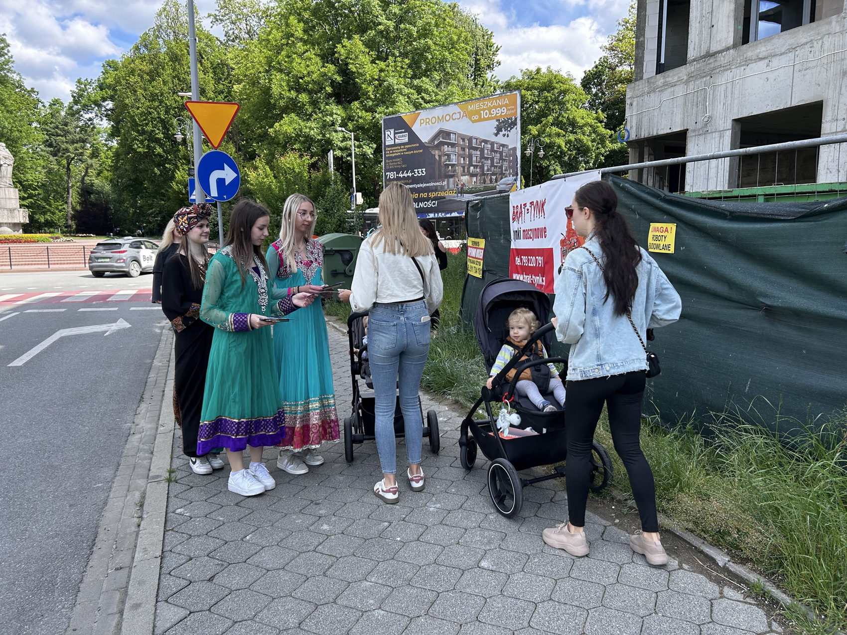 Happening młodzieżowy – SDL (3) Ulica, chodnik. Trzy dziewczyny w kolorowych indyjskich sukienkach stoją na chodniku i rozdają ulotki mijanym kobietom z wózkami. Po prawej stronie widać betonową konstrukcję budynku.