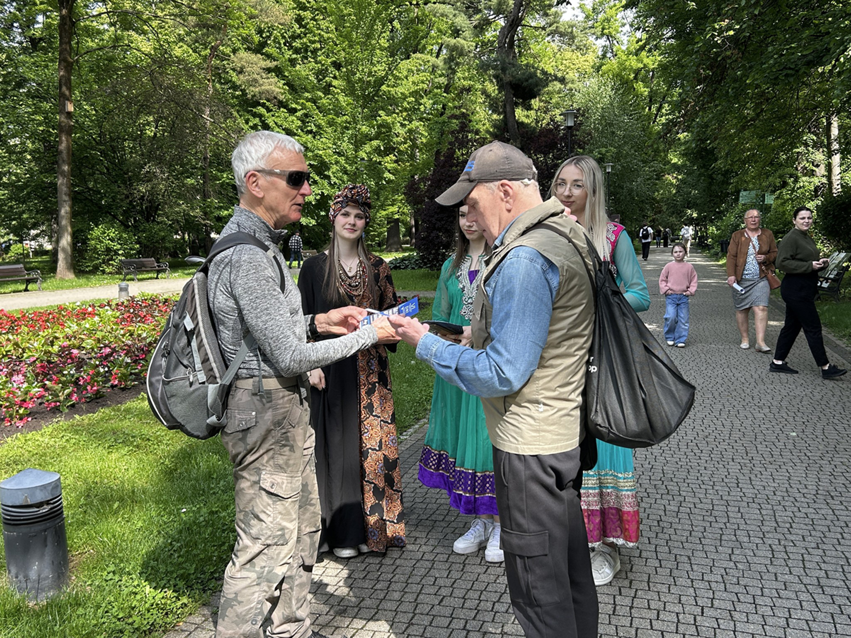 Happening młodzieżowy – SDL (4) Na zdjęciu, w parku, widać grupę ludzi. Po lewej stronie stoi mężczyzna w okularach przeciwsłonecznych, szarej koszulce z długim rękawem i spodniach moro, z plecakiem na plecach. Wręcza coś mężczyźnie stojącemu naprzeciwko. Mężczyzna po prawej stronie ma na sobie dżinsową koszulę, beżową kamizelkę i czapkę z daszkiem. Za nim stoją trzy młode kobiety ubrane w tradycyjne zagraniczne, kolorowe stroje. Jedna z nich ma na głowie ozdobny turban. W tle widać park z zielonymi drzewami, kwiatami i ławkami. Na ścieżce w oddali widać inne osoby spacerujące po parku.