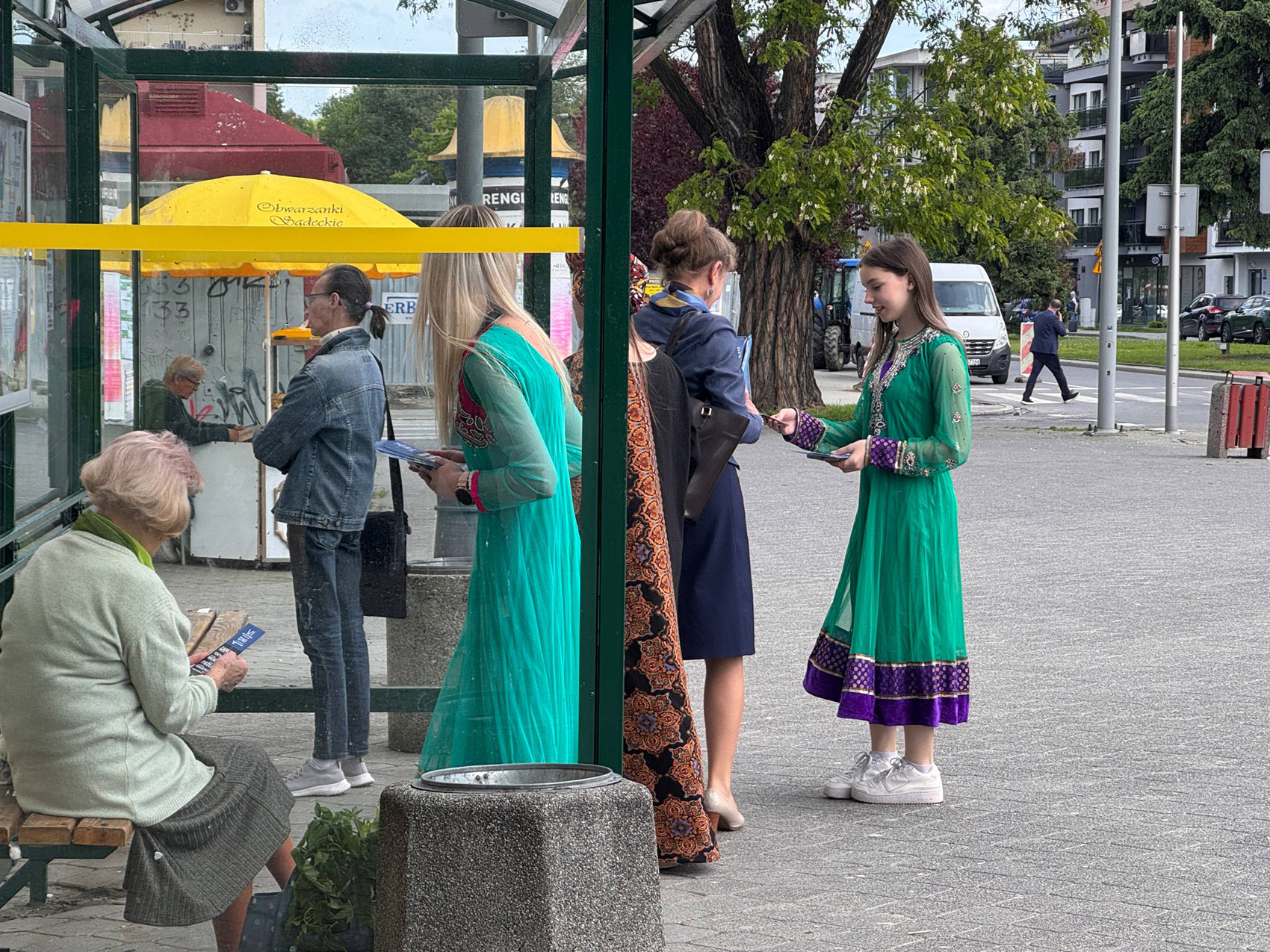 Happening młodzieżowy – SDL (5) Na zdjęciu widać grupę osób w przestrzeni miejskiej obok przystanku autobusowego. Na pierwszym planie, po lewej stronie, siedzi starsza kobieta w zielonym swetrze i szarej spódnicy, czytająca ulotkę. Na pierwszym planie, po prawej stronie, stoi młoda dziewczyna w zielonej sukience z fioletowymi zdobieniami, wręczająca ulotkę kobiecie w granatowej sukience. Obok nich stoi kobieta w turkusowej sukience. W tle widać ulicę z przechodzącym mężczyzną w garniturze, zaparkowane samochody i budynki.