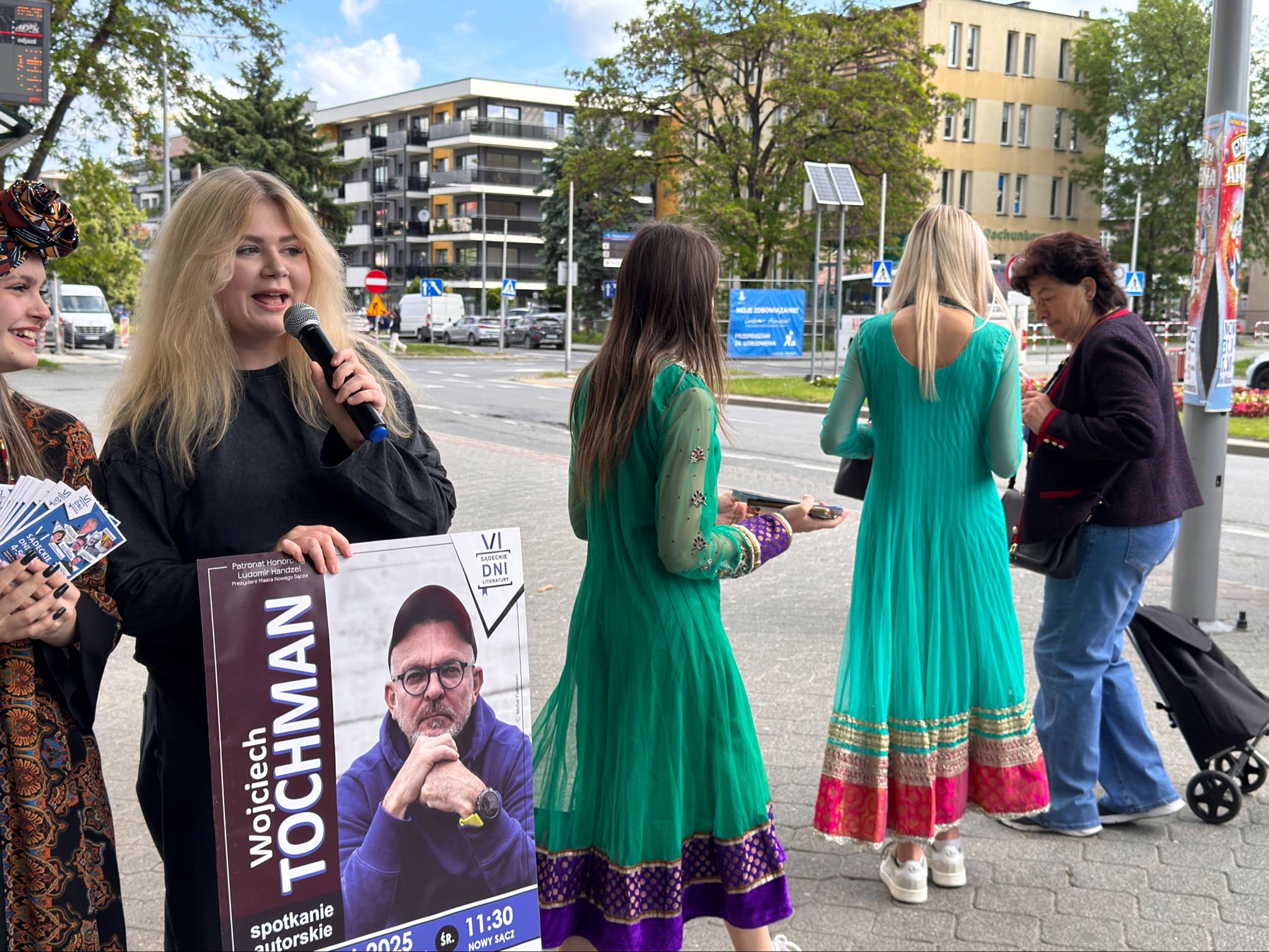 Happening młodzieżowy – SDL (6) Na zdjęciu w plenerze widać grupę osób. Z przodu po lewej stronie stoi młoda kobieta z blond włosami, trzymająca mikrofon i plakat z wizerunkiem Wojciecha Tochmana oraz informacją o spotkaniu autorskim. Obok niej stoi dziewczyna z ulotkami w ręku i chustą na głowie. W tle widać dwie kobiety w długich, kolorowych sukniach, a za nimi starszą kobietę z torbą na kółkach. W tle znajdują się budynki miejskie i ulica.