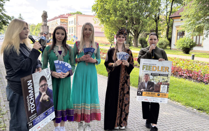 Na zdjęciu stoi pięć młodych kobiet. Dwie z nich trzymają mikrofony przy twarzy. Pozostałe trzy kobiety ubrane są w tradycyjne stroje, prawdopodobnie reprezentujące różne kultury. Każda z nich trzyma w rękach plik niebieskich ulotek. Po lewej stronie, kobieta z mikrofonem trzyma plakat z wizerunkiem Wojciecha Tochmana i informacją o spotkaniu autorskim 4 czerwca 2025 roku o godzinie 11:30 w Parku im. Ireny Styczyńskiej w Nowym Sączu. Po prawej stronie, kobieta z mikrofonem trzyma plakat z wizerunkiem Arkadego Radosława Fiedlera i informacją o spotkaniu autorskim 5 czerwca 2025 roku o godzinie 13:00 w tym samym parku. Plakaty informują również o "VI Sądeckich Dniach".
