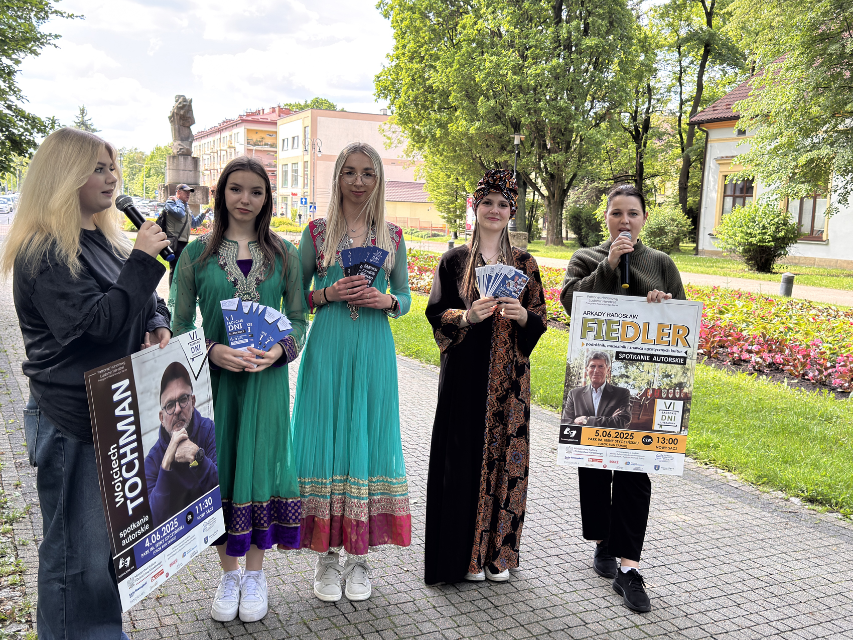 Happening młodzieżowy – SDL (7) Na zdjęciu stoi pięć młodych kobiet. Dwie z nich trzymają mikrofony przy twarzy. Pozostałe trzy kobiety ubrane są w tradycyjne stroje, prawdopodobnie reprezentujące różne kultury. Każda z nich trzyma w rękach plik niebieskich ulotek. Po lewej stronie, kobieta z mikrofonem trzyma plakat z wizerunkiem Wojciecha Tochmana i informacją o spotkaniu autorskim 4 czerwca 2025 roku o godzinie 11:30 w Parku im. Ireny Styczyńskiej w Nowym Sączu. Po prawej stronie, kobieta z mikrofonem trzyma plakat z wizerunkiem Arkadego Radosława Fiedlera i informacją o spotkaniu autorskim 5 czerwca 2025 roku o godzinie 13:00 w tym samym parku. Plakaty informują również o "VI Sądeckich Dniach".