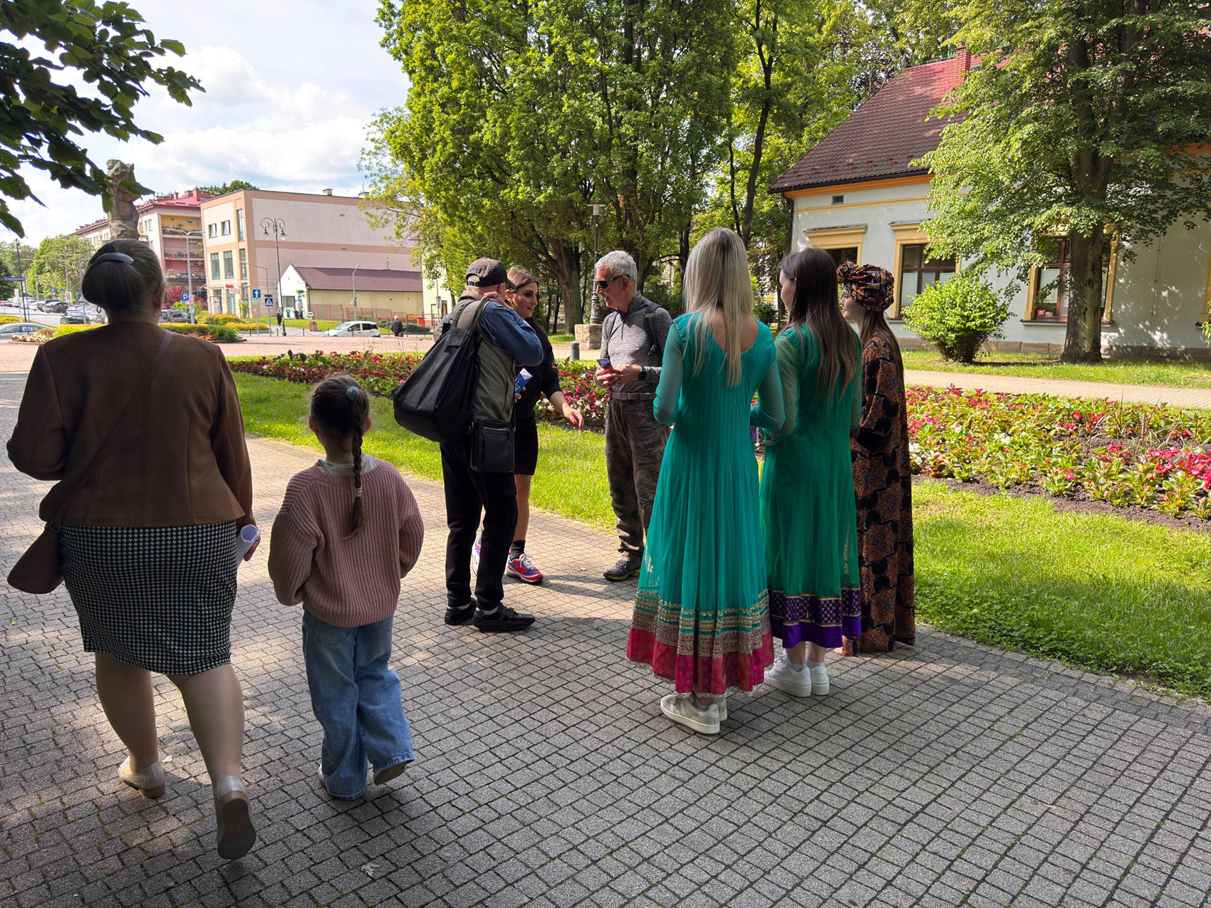 Happening młodzieżowy – SDL (9) Na zdjęciu widać grupę ludzi w parku. Po lewej stronie kadru idzie kobieta w brązowej kurtce i spódnicy w pepitkę, a obok niej dziewczynka w różowym swetrze i dżinsach. W centrum zdjęcia stoi grupa osób, w tym dwie kobiety w długich, turkusowych sukniach z kolorowymi zdobieniami. Obok nich stoi kobieta w długiej, wzorzystej sukni i nakryciu głowy. Mężczyźni w grupie rozmawiają ze sobą. W tle widać budynki i drzewa, a także klomby z kwiatami.