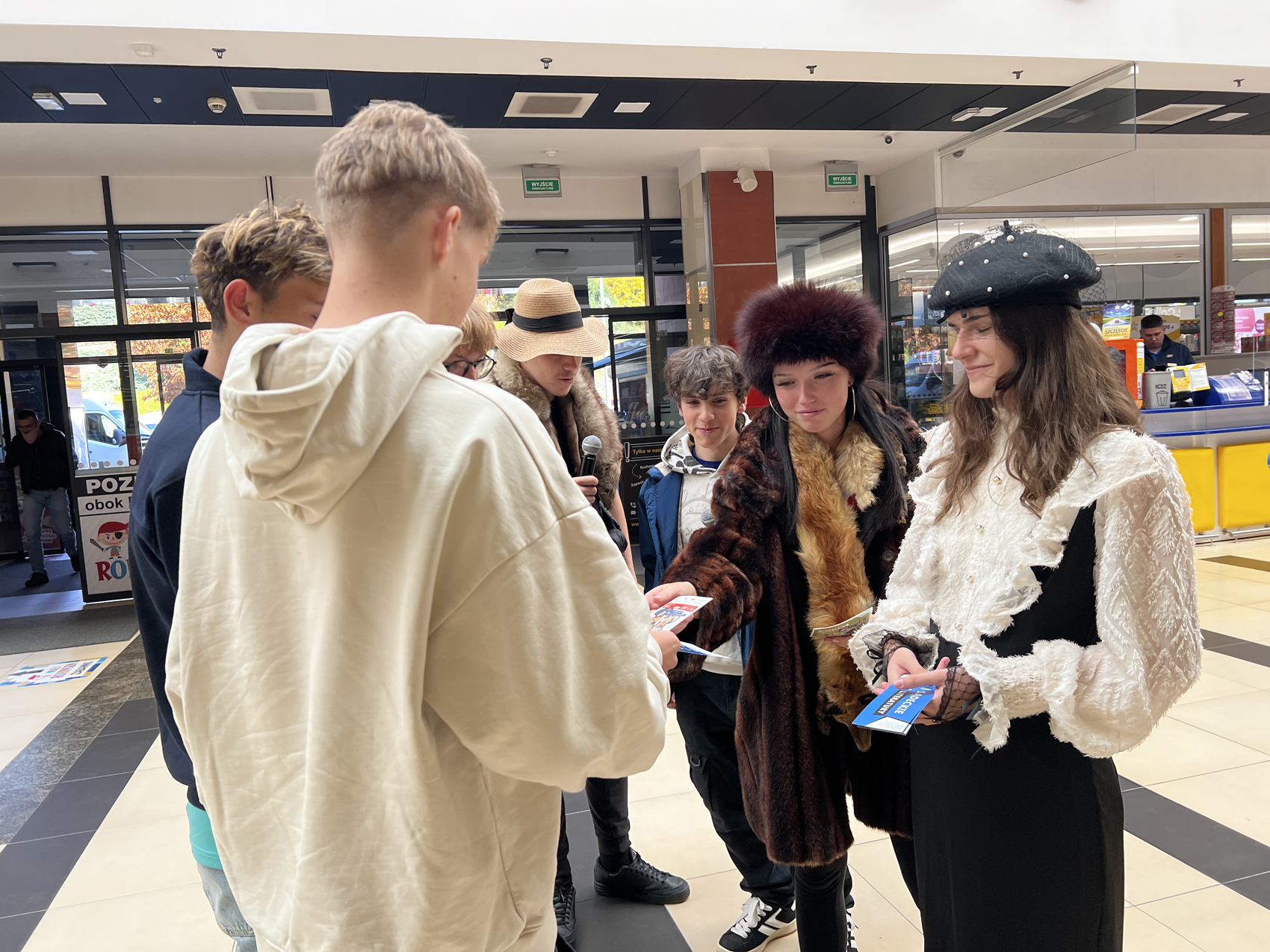 Osiem osób w pomieszczeniu. W tle kiosk Lotto. Cztery młode osoby w przebraniach: po lewej chłopak w dużym słomianym kapeluszu i z szalem lisem na szyi. Trzyma mikrofon w ręce. Obok niego stoi chłopak w czarnych spodniach, szarej bluzie i niebieskiej kurtce. Trzecia z osób, to dziewczyna ubrana w grube ciemnobrązowe futro i futrzaną czapkę do kompletu. Na szyi także ma lisa. Nachyla się do przodu i podaje ulotkę chłopaki, który stoi przed nią. Po prawej natomiast stoi druga dziewczyna w długich brązowych włosach, ubrana w długą czarną sukienkę z białą koszulą z dużymi falbanami. Na głowie ma czarny beret z siateczką i perełkami. W ręce trzyma ulotki. Na przeciwko nich stoi trzech młodych chłopaków. Stoją tyłem. Jeden z nich w rękach trzyma ulotki.