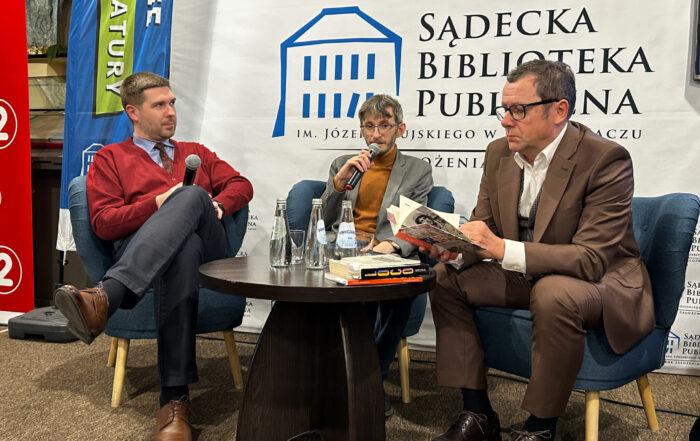 W pomieszczeniu na środku na trzech fotelach siedzi trzech mężczyzn. Przed nimi na środku stoi brązowy okrągły stolik. Na stoliku znajdują się: trzy butelki z wodą, trzy szklanki, kilka książek. Mężczyzna pierwszy od prawej strony ubrany jest w brązowy garnitur. Trzyma w rękach otwartą książkę, czyta. Mężczyzna na środku ubrany jest w szarą marynarkę i musztardowy golf. Trzyma mikrofon blisko ust i mówi do niego. Mężczyzna pierwszy od lewej strony ubrany jest w bordowy sweter i ciemne spodnie. Patrzy do góry. W tle za siedzącymi mężczyznami stoi duża biała ścianka z logotypem Sądeckiej Biblioteki Publicznej.
