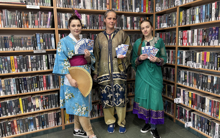 Trzy dziewczyny stoją na tle wysokich regałów z książkami. Po prawej stoi dziewczyna w niebieskich szlafroku w białe kwiaty. Przewiązana jest fioletowym pasem. Ma koczka na głowie i wpięte we włosy fioletowe kwiaty. W jednej ręce trzyma drewniany wachlarz, a w drugiej niebieskie ulotki. Obok niej stoi druga kobieta w związanych włosach w koczek. Ma na sobie jasny strój z granatowo złotą narzutą. W ręce również trzyma kolorowe ulotki. Trzecia z kobiet ma na sobie długa zieloną sukienkę w stylu indyjskim z złoto-fioletowymi wstawkami. Ma włosy związane w warkocz. W rękach również trzyma ulotki.