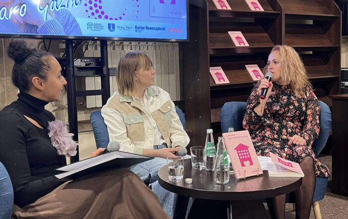 Na zdjęciu widać trzy kobiety siedzące na niskich niebieskich fotelach. W tle widać fragment ekranu z nazwiskiem "Karo Gazik" i innymi informacjami, a także półki z książkami. Kobieta po prawej stronie trzyma mikrofon i mówi, ubrana jest w kwiecistą sukienkę. Dwie pozostałe kobiety siedzą obok niej i słuchają. Na stole przed nimi stoją butelki z wodą, szklanki i książki.