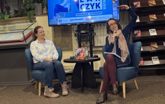 Dwie kobiety siedzą na niskich niebieskich fotelach. Kobieta po lewej uśmiecha się i patrzy na drugą z kobiet. Ma na sobie jasną wzorzystą koszulę i jeansy. Druga z kobiet ma krótkie ciemne włosy, ciemną sukienkę i jasny szalik. Między nimi stoi niski ciemny stolik, na którym stoi książka i szklane butelki wody oraz regał z książkami. Z nimi znajduje się duży ekran z niebieską grafiką i regał z książkami.