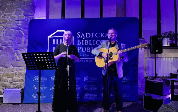 Na scenie, oświetlonej niebieskim światłem, występuje duet muzyczny. Kobieta w długiej, czarnej sukni stoi po lewej stronie, śpiewając do mikrofonu. Obok niej, po prawej, mężczyzna gra na akustycznej gitarze, również śpiewając do mikrofonu. W tle widoczny jest baner z napisem "Sądecka Biblioteka Publiczna". Za nimi znajdują się okna, a po prawej stronie kolumna. Na scenie widoczne są również statywy mikrofonowe, nuty, głośniki i wzmacniacze. Atmosfera jest intymna i skupiona na występie.