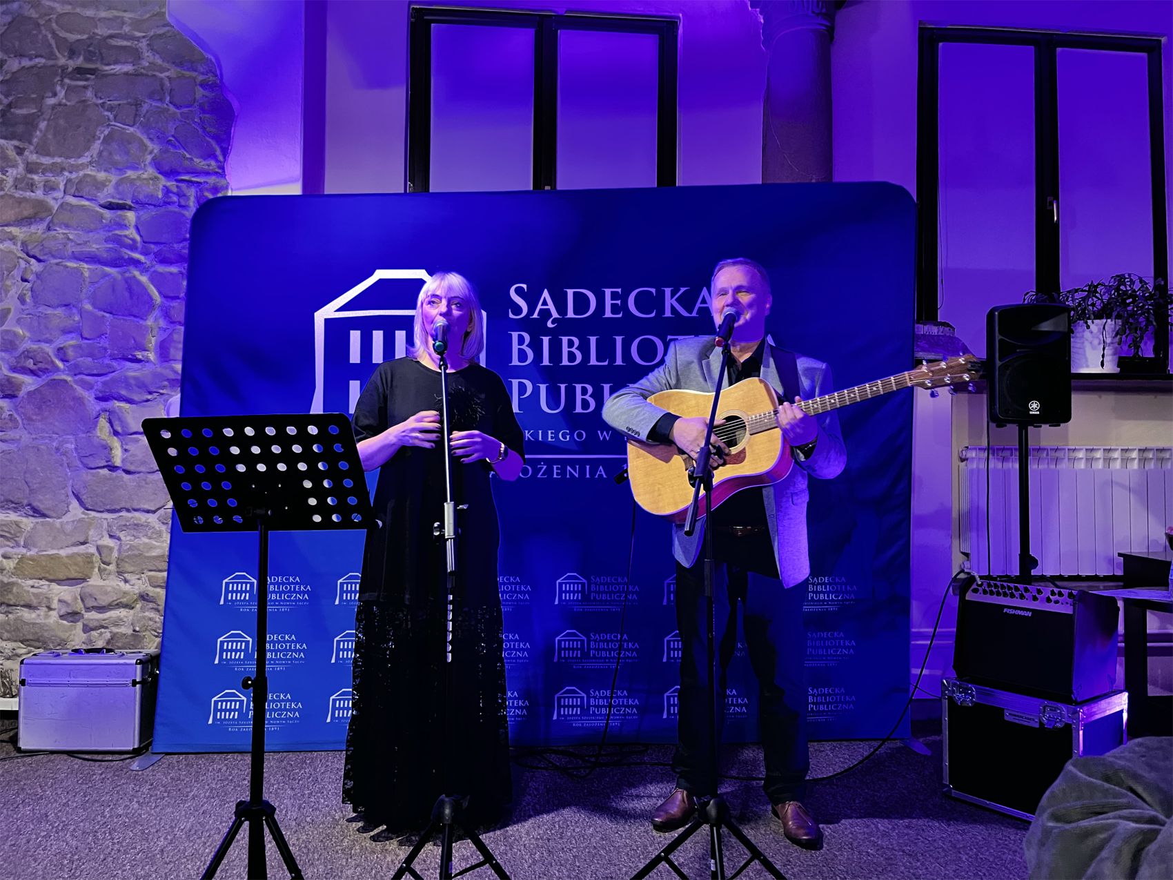 Na scenie, oświetlonej niebieskim światłem, występuje duet muzyczny. Kobieta w długiej, czarnej sukni stoi po lewej stronie, śpiewając do mikrofonu. Obok niej, po prawej, mężczyzna gra na akustycznej gitarze, również śpiewając do mikrofonu. W tle widoczny jest baner z napisem "Sądecka Biblioteka Publiczna". Za nimi znajdują się okna, a po prawej stronie kolumna. Na scenie widoczne są również statywy mikrofonowe, nuty, głośniki i wzmacniacze. Atmosfera jest intymna i skupiona na występie.