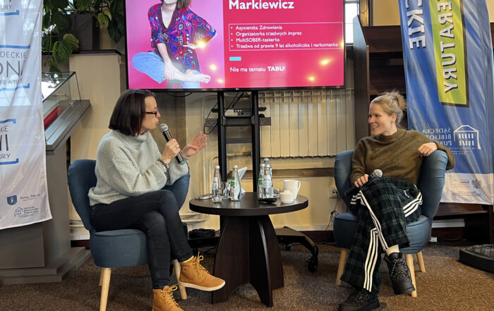 Dwie kobiety siedzą na niskich niebieskich fotelach. Kobieta po lewej ma krótkie brązowe włosy. Ubrana jest w szary sweterek, ciemne jeansy i żółte buty. Trzyma w ręce mikrofon i mówi do niego. Druga z kobiet uśmiecha się. Ma jasne, spięte włosy. Ubrana jest w ciemnozielony sweter i ciemne spodnie w kratę. MA czarne buty. Trzyma w ręce mikron. Pomiędzy nimi znajduje się niski ciemny stolik. Na stoliku stoją szklane butelki i filiżanka. Nad nimi widać duży ekran z prezentacją. Na zdjęciu kobieta w kolorowym ubraniu i podpis: Marta Markiewicz.